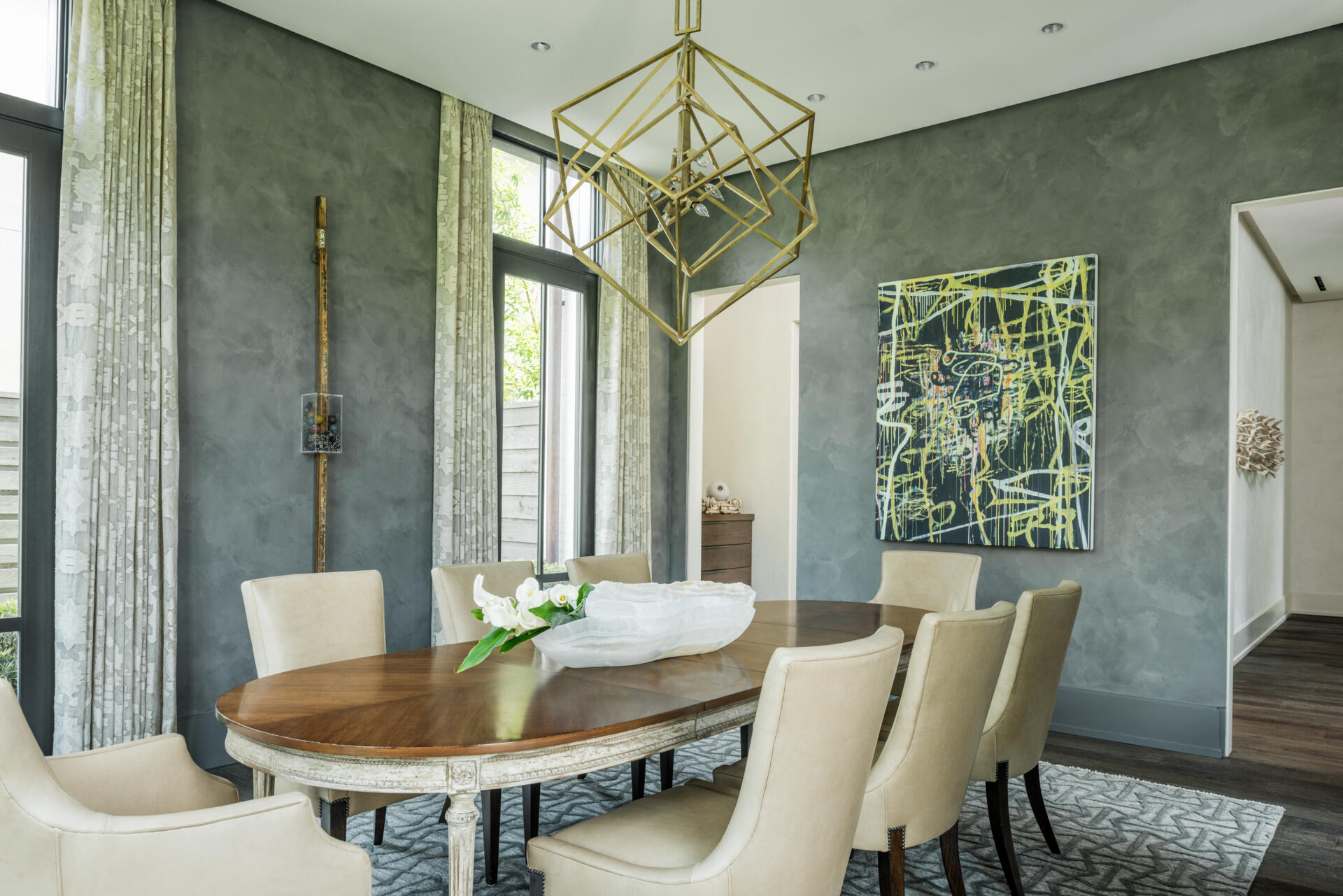A formal dining room featuring charcoal-gray textured walls, an oval wood dining table, cream upholstered chairs, colorful abstract art, and a sculptural gold geometric chandelier.