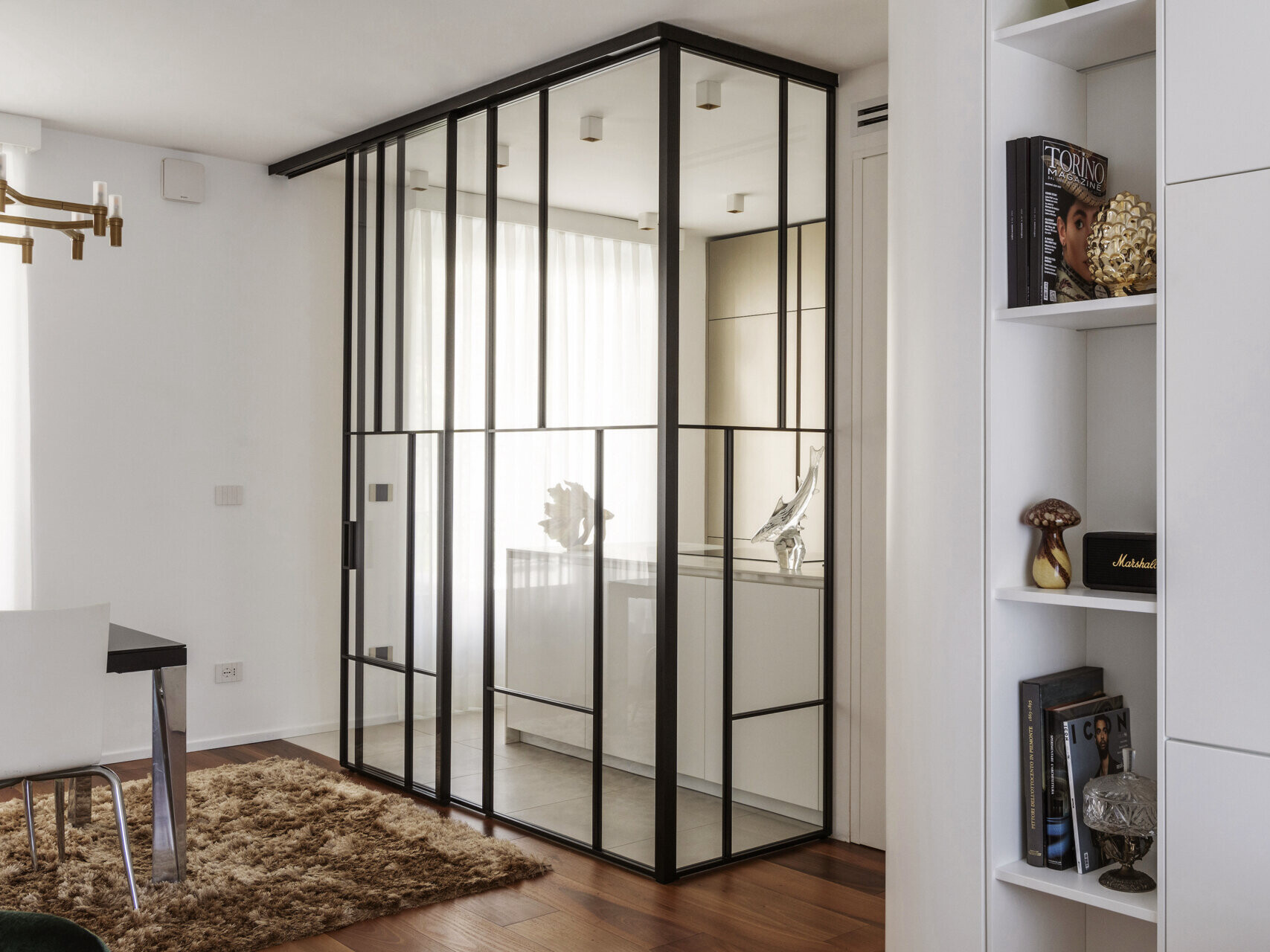 A glass-enclosed kitchen in a Turin apartment features floor-to-ceiling black metal frames, a sliding glass door, minimalist cabinetry, and marble-look countertops.