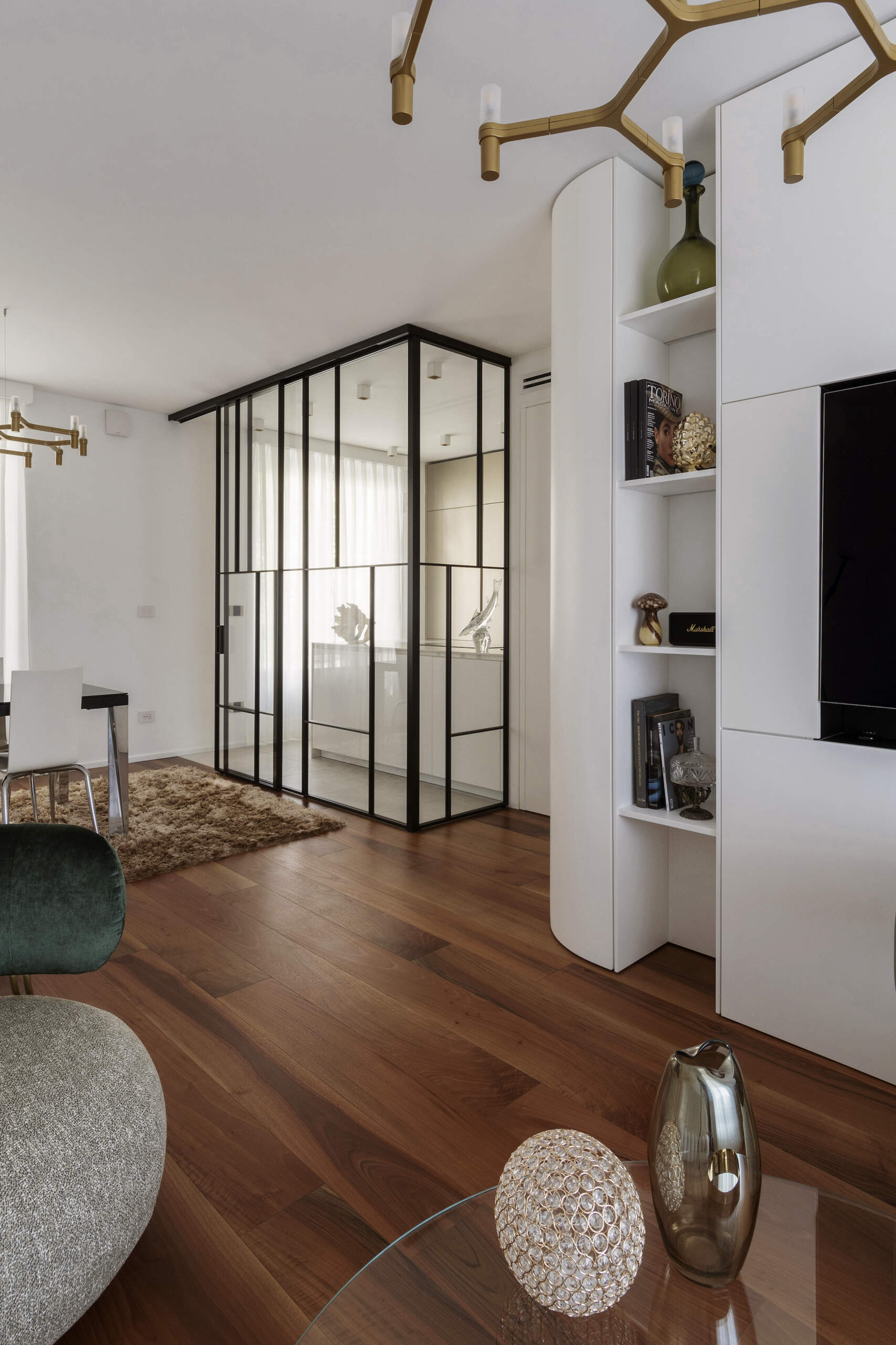 A glass-enclosed kitchen in a Turin apartment features floor-to-ceiling black metal frames, a sliding glass door, minimalist cabinetry, and marble-look countertops.