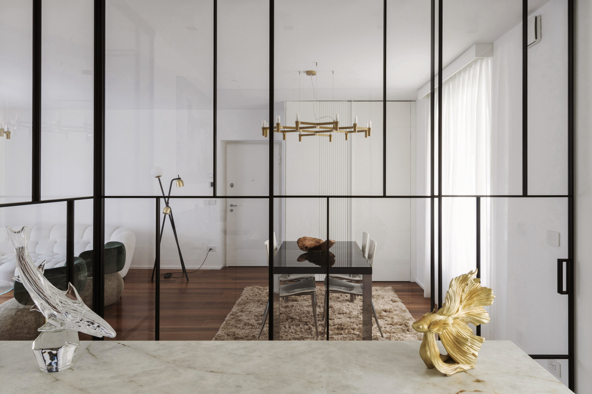 A glass-enclosed kitchen in a Turin apartment features floor-to-ceiling black metal frames, a sliding glass door, minimalist cabinetry, and marble-look countertops.