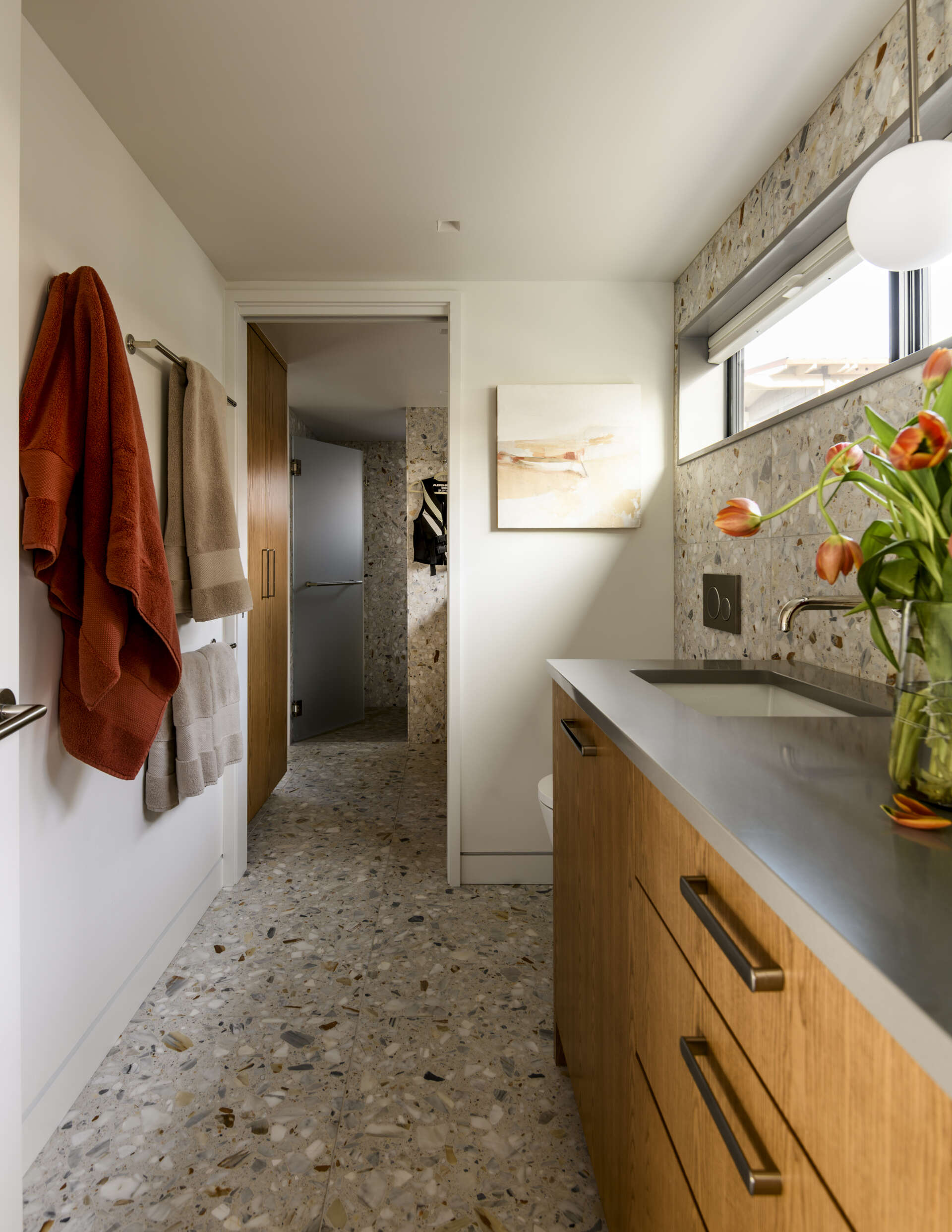 Featuring terrazzo-style floor and wall tiles, warm wood cabinetry, a gray countertop, wall-mounted faucet, and a white globe pendant light, this bathroom combines natural textures with contemporary design.