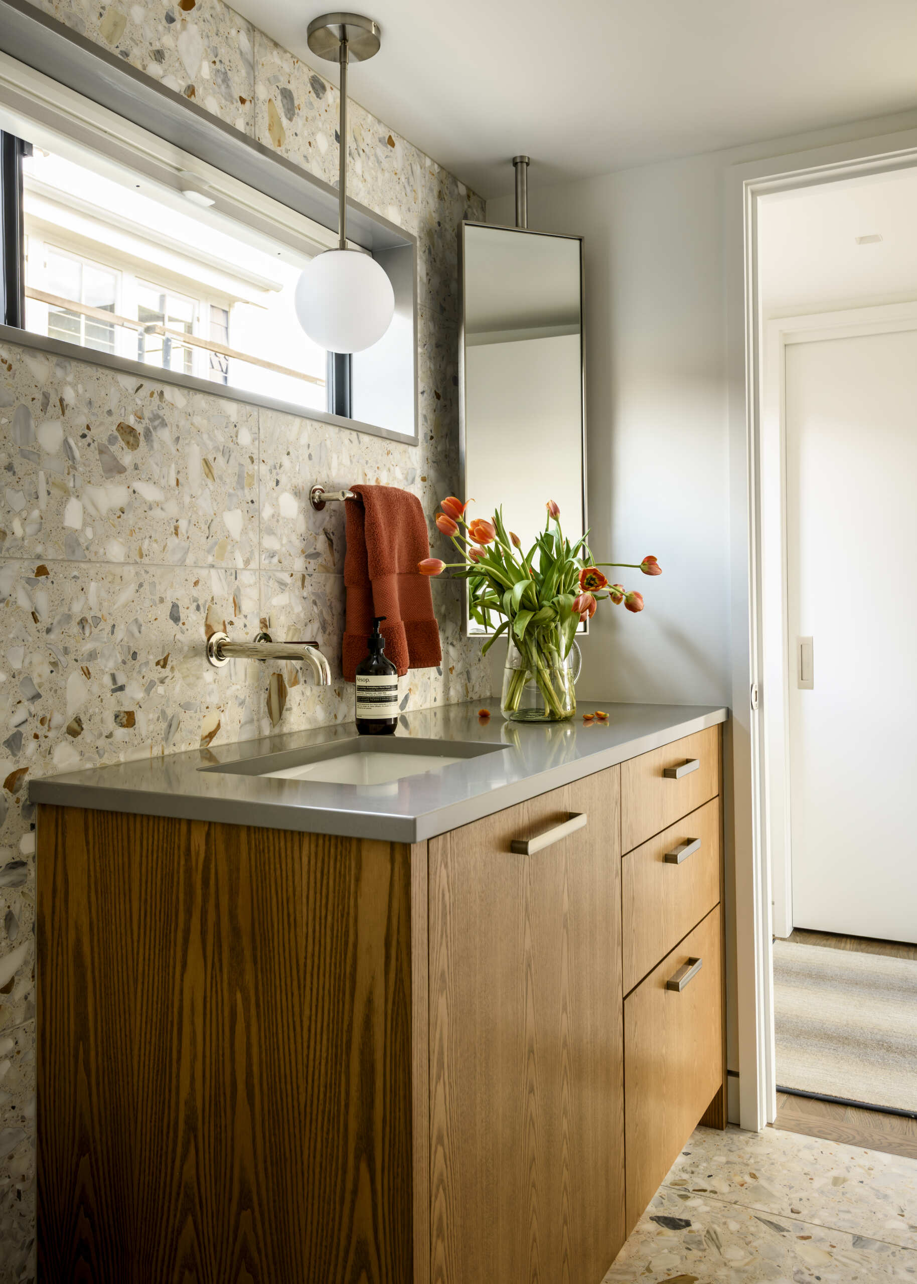 Featuring terrazzo-style floor and wall tiles, warm wood cabinetry, a gray countertop, wall-mounted faucet, and a white globe pendant light, this bathroom combines natural textures with contemporary design.