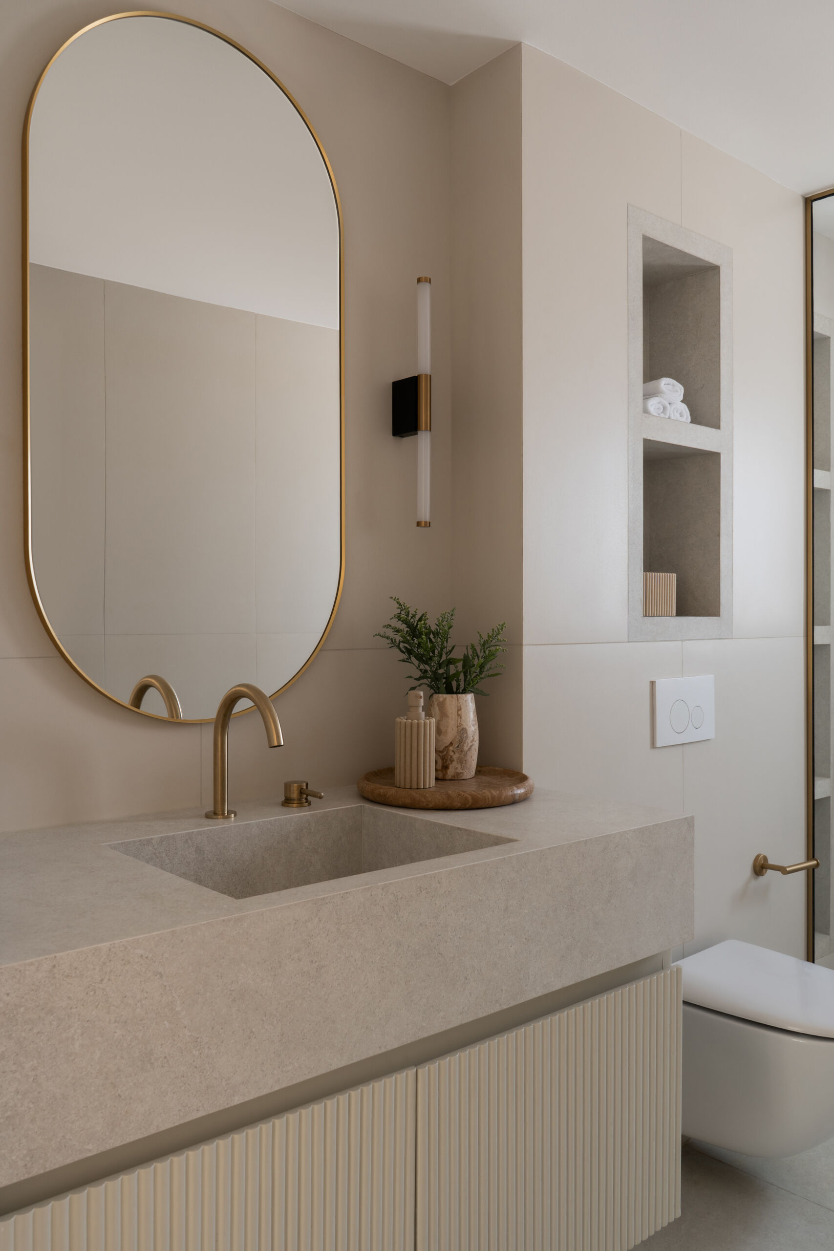 A contemporary guest bathroom in The White Arcade, Dubai, featuring a natural stone vanity with an integrated sink, arched brass-framed mirror, ribbed cabinetry, walk-in glass shower, and beautiful brass fixtures. Designed by D’Ora Tokai Designs.