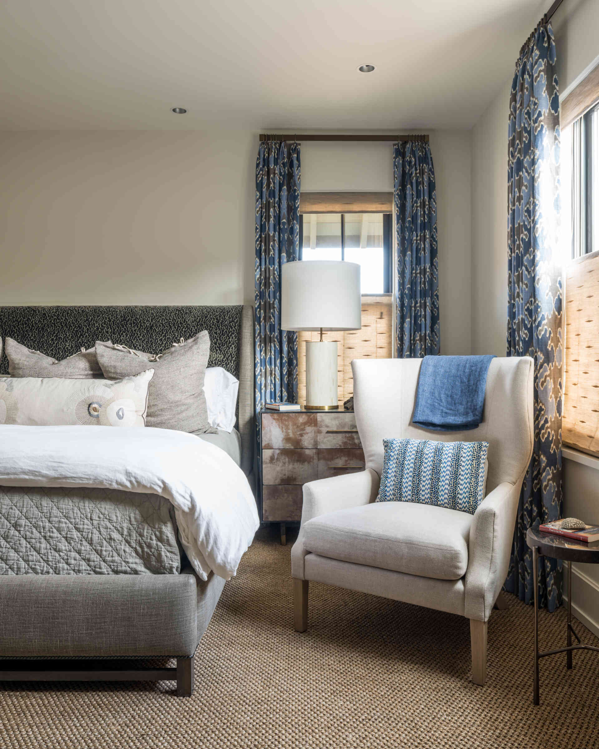 A neutral guest bedroom with an upholstered bed, blue patterned curtains, woven Roman shades, cream wingback chair, metallic bedside cabinet, and textured carpet.