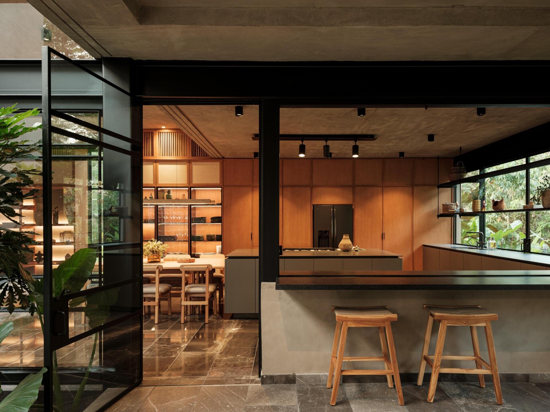 A modern indoor-outdoor home bar in Mexico features a dark stone countertop, wooden bar stools, and large glass openings that connect the kitchen to a covered terrace with a fireplace.