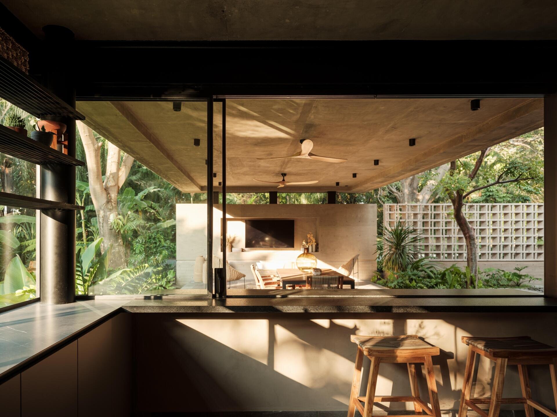 A modern indoor-outdoor home bar in Mexico features a dark stone countertop, wooden bar stools, and large glass openings that connect the kitchen to a covered terrace with a fireplace.