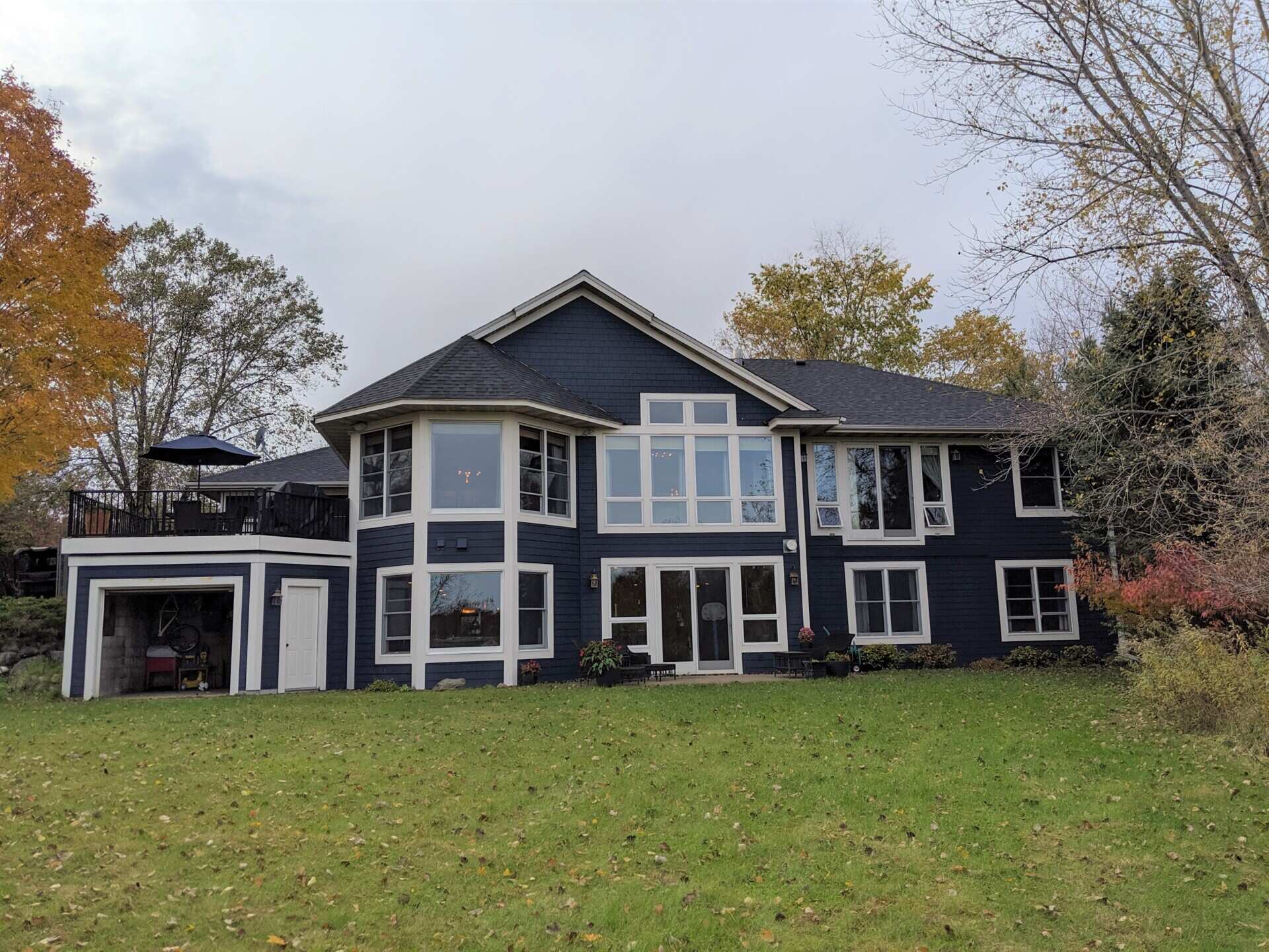 Rear view of a traditional 1990s house before renovation, showing blue siding, white-framed windows, pitched roofs, and an elevated outdoor deck overlooking the backyard.