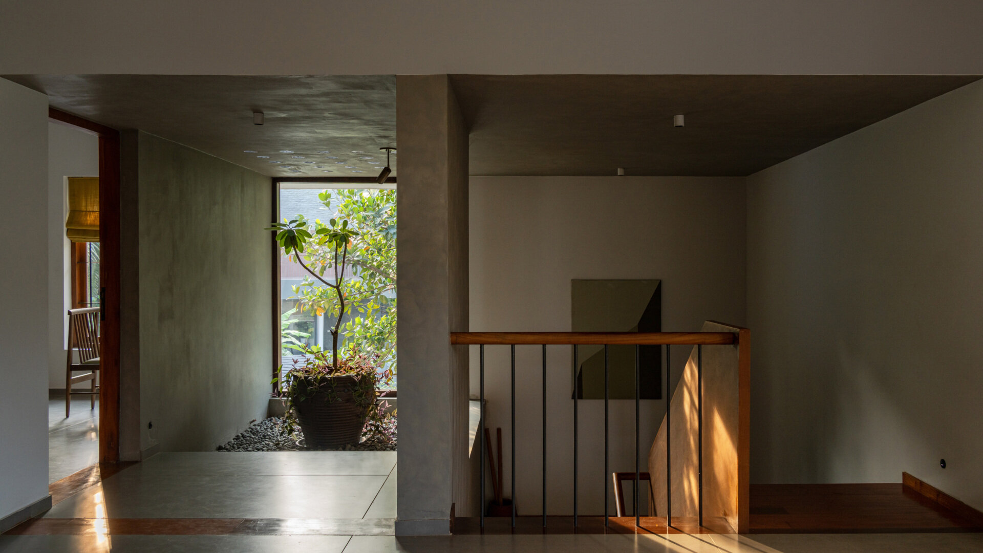 A small indoor courtyard in a Kerala home features a potted tree, pebble-covered floor, natural ventilation, and sunlight creating beautiful shadows on the walls.