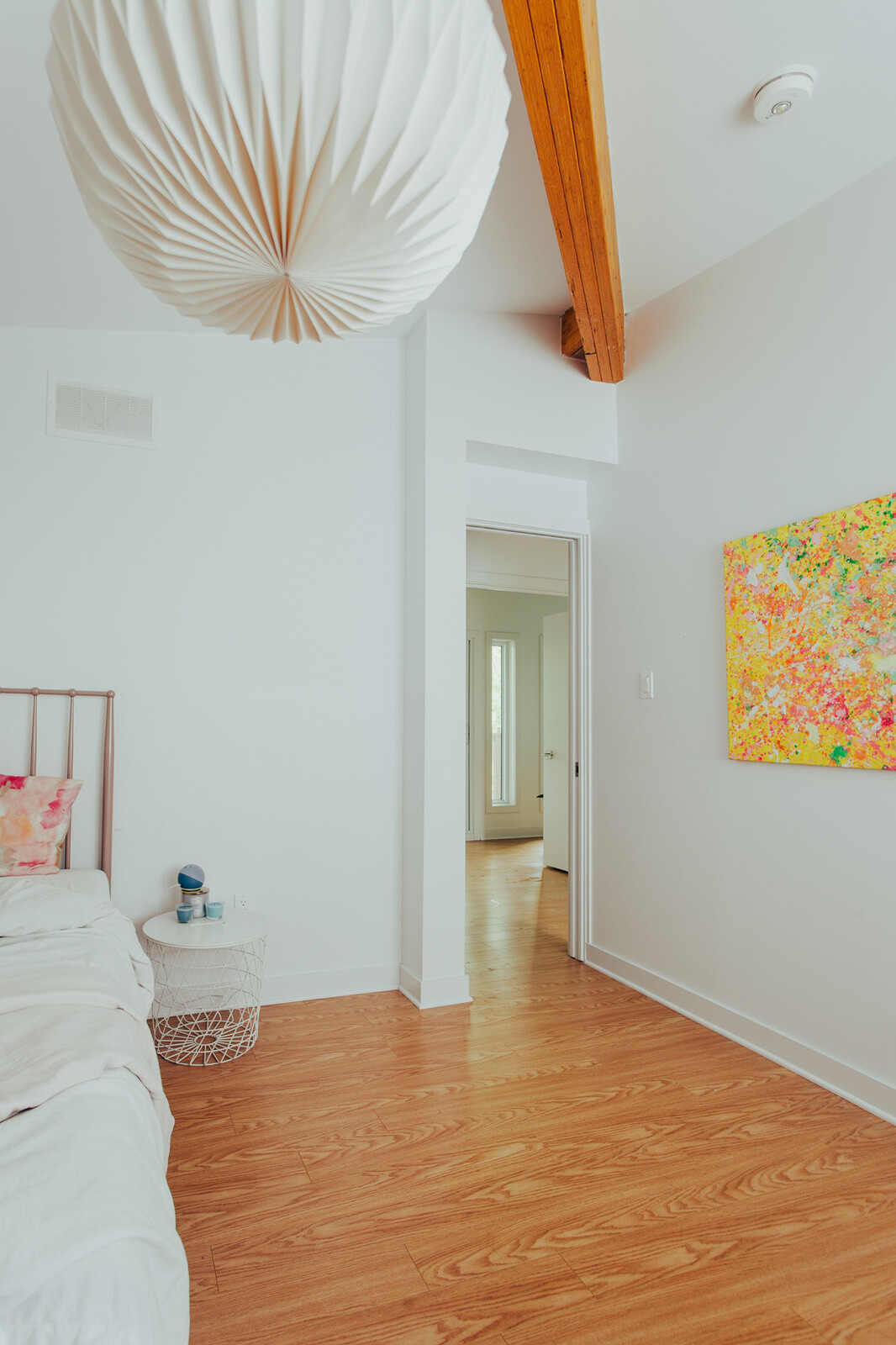 Explore a bright upstairs bedroom with white walls, warm wooden floors, an exposed ceiling beam, colorful artwork, and space-saving pocket doors in a renovated family home.