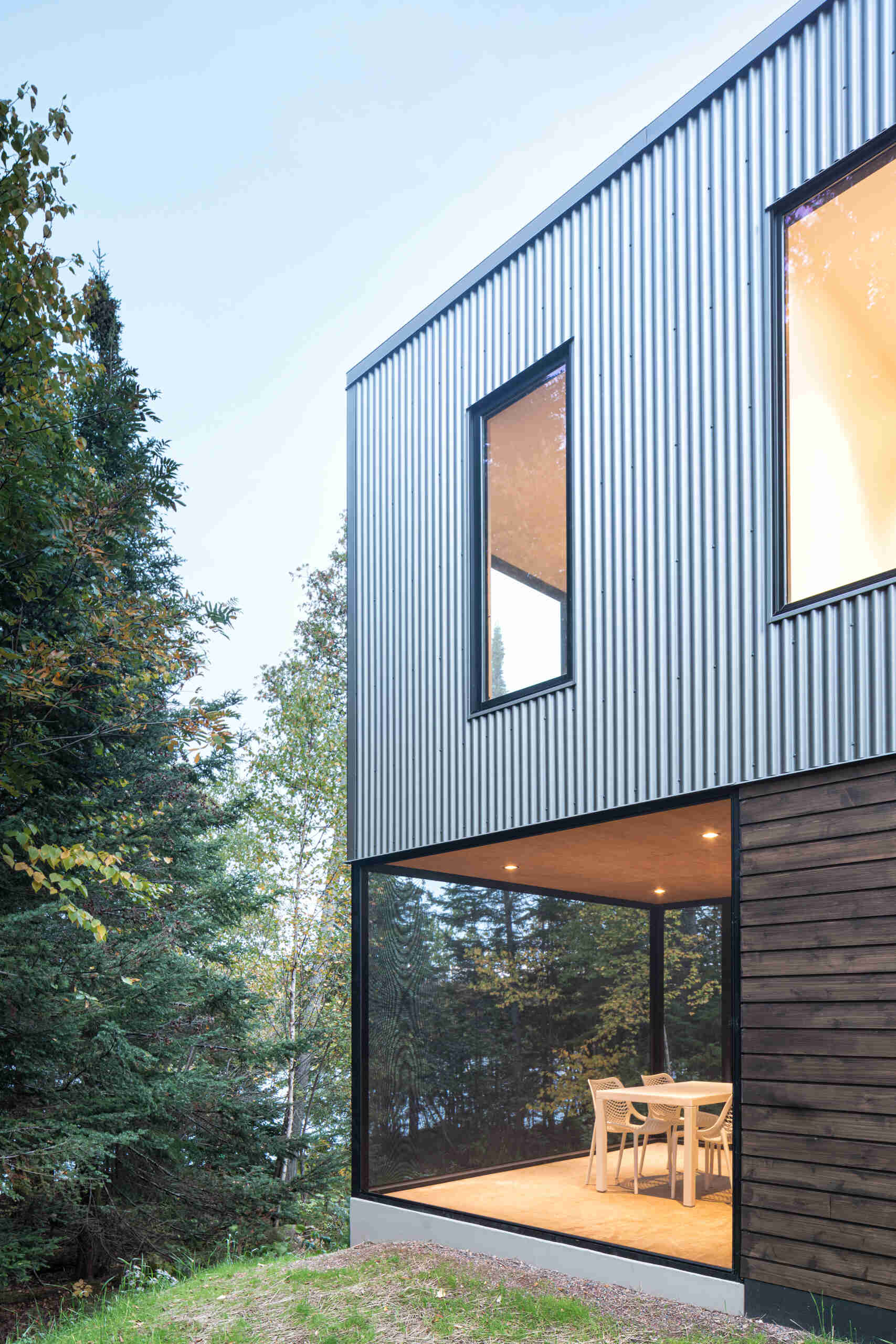 A screened porch beneath the cantilevered upper floor of a modern cabin, furnished with a white outdoor dining table and chairs. Black mesh screens enclose the space while preserving views of the surrounding woodland.