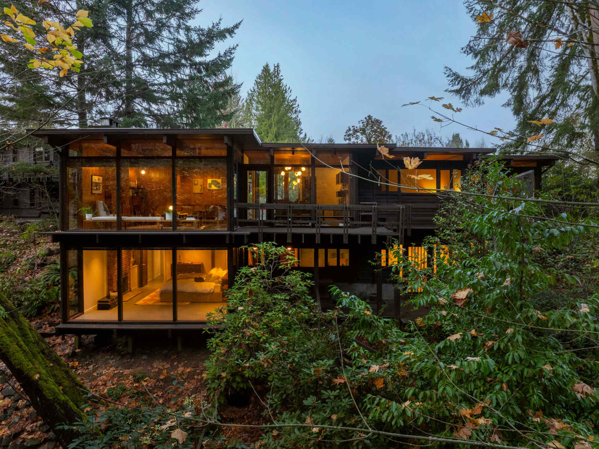 Explore the architecture of The Burrows in Seattle, featuring dark timber siding, expansive glass walls, elevated wooden decking, and two levels overlooking a forested ravine.