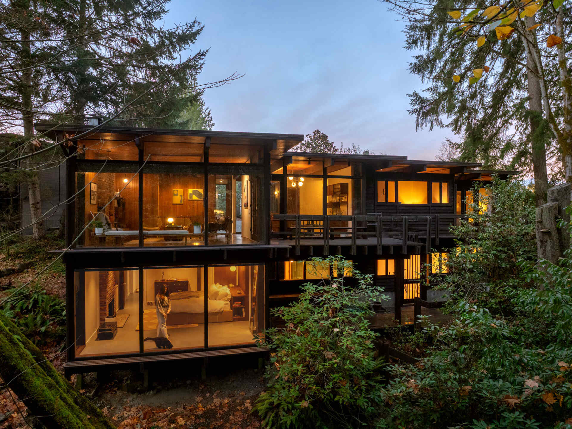 Explore the architecture of The Burrows in Seattle, featuring dark timber siding, expansive glass walls, elevated wooden decking, and two levels overlooking a forested ravine.