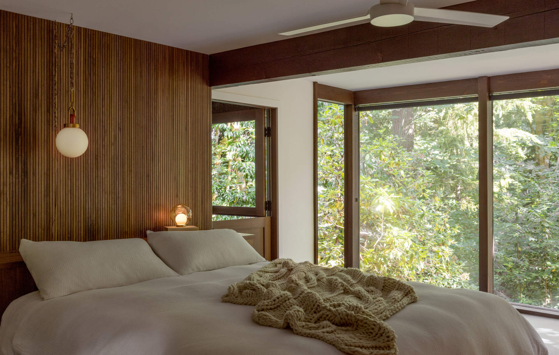Explore the primary bedroom of The Burrows, a renovated mid-century home in Seattle by chadbourne + doss architects. A custom wooden bed and slatted headboard, original brick fireplace, neutral bedding, and floor-to-ceiling windows overlooking the forest create a warm, inviting space.