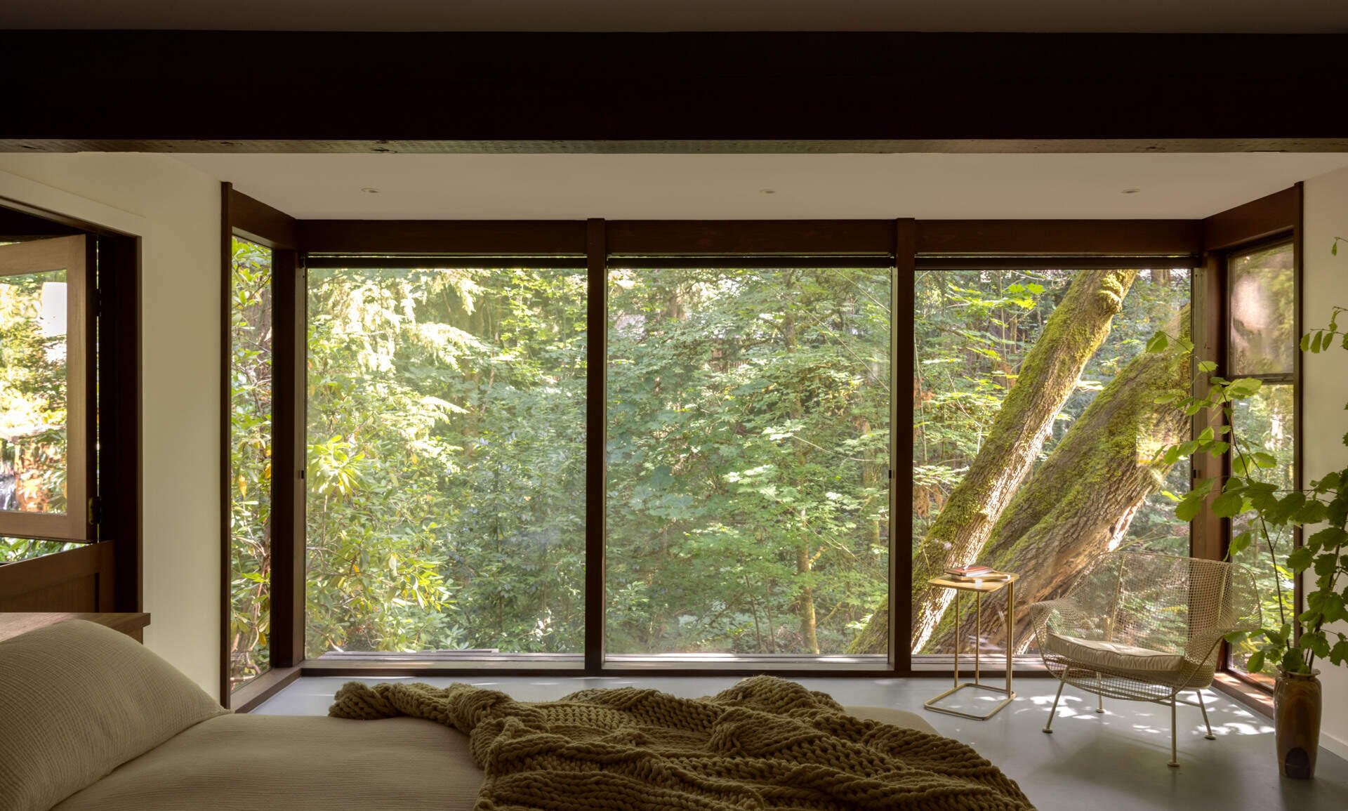 Explore the primary bedroom of The Burrows, a renovated mid-century home in Seattle by chadbourne + doss architects. A custom wooden bed and slatted headboard, original brick fireplace, neutral bedding, and floor-to-ceiling windows overlooking the forest create a warm, inviting space.