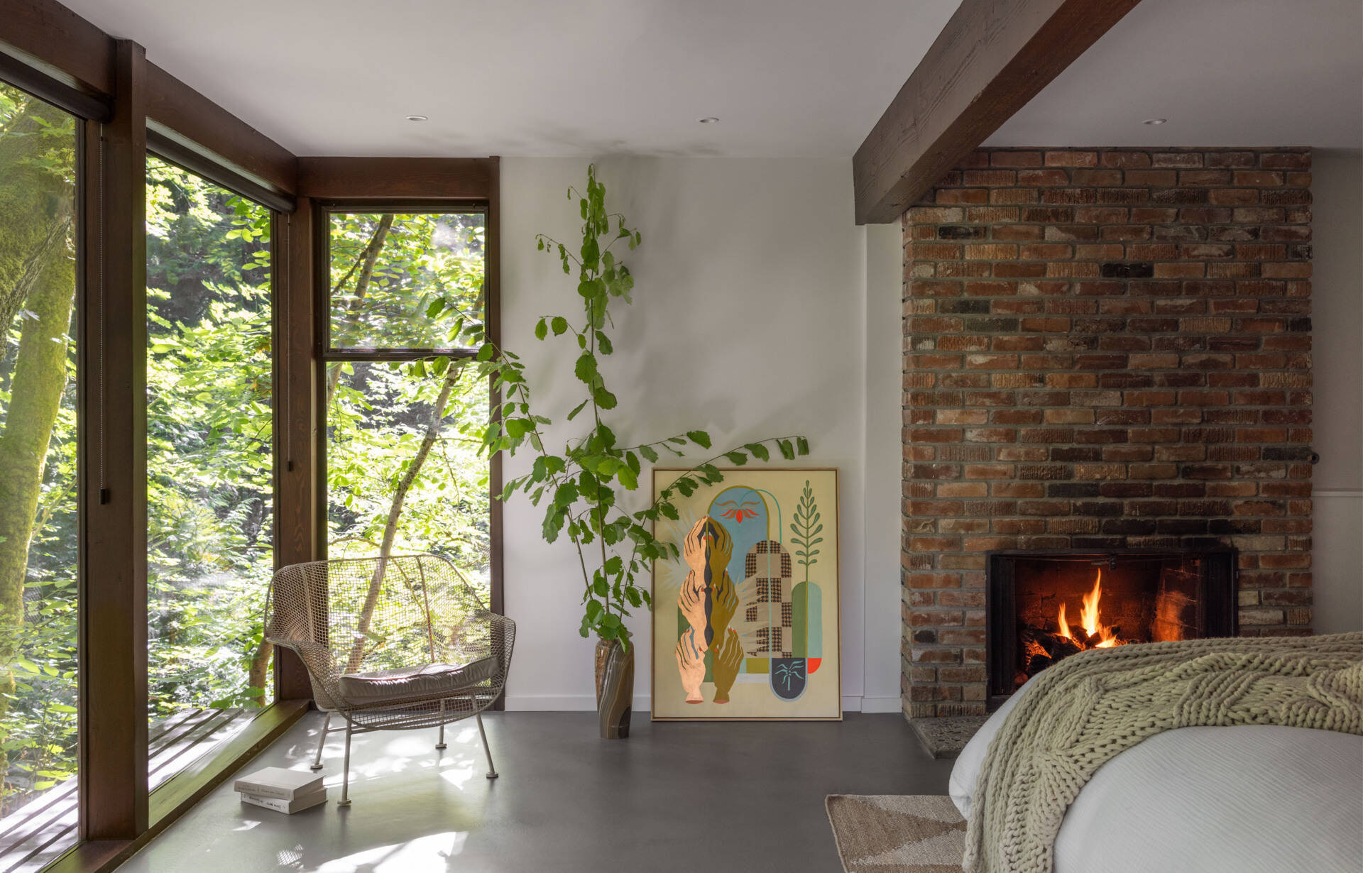 Explore the primary bedroom of The Burrows, a renovated mid-century home in Seattle by chadbourne + doss architects. A custom wooden bed and slatted headboard, original brick fireplace, neutral bedding, and floor-to-ceiling windows overlooking the forest create a warm, inviting space.