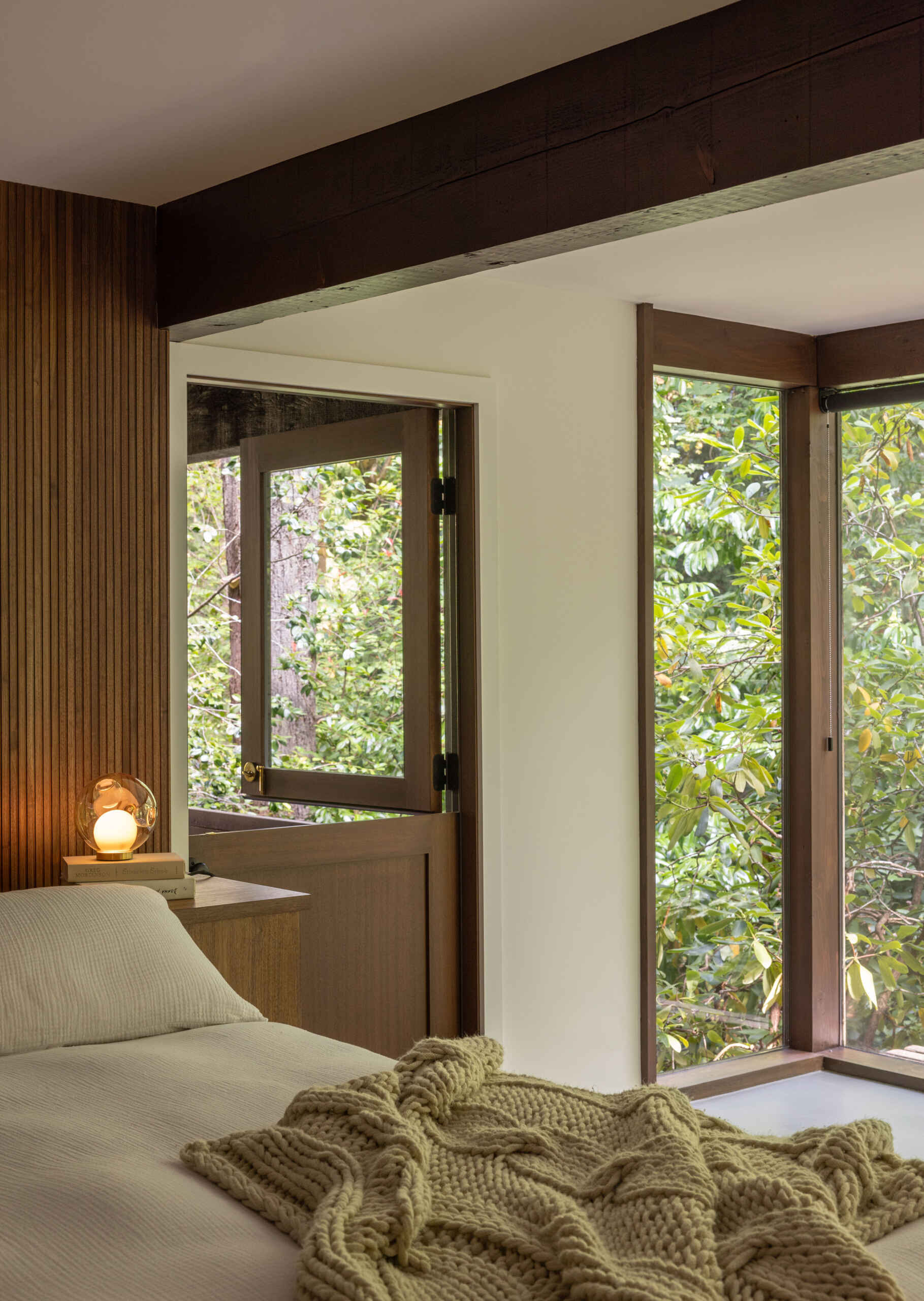 Explore the primary bedroom of The Burrows, a renovated mid-century home in Seattle by chadbourne + doss architects. A custom wooden bed and slatted headboard, original brick fireplace, neutral bedding, and floor-to-ceiling windows overlooking the forest create a warm, inviting space.
