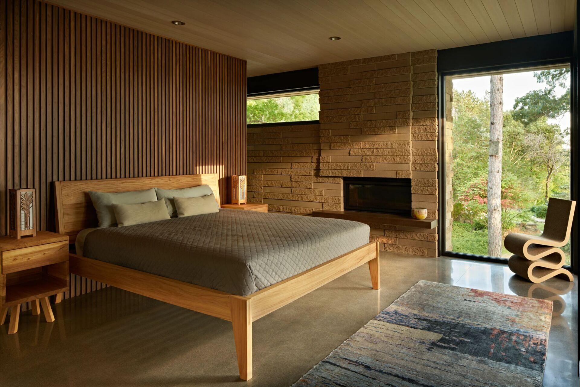 A modern bedroom with a wooden platform bed, vertical slatted wood accent wall, textured masonry fireplace, and polished concrete flooring. A sculptural curved chair sits beside a large corner window overlooking the garden.