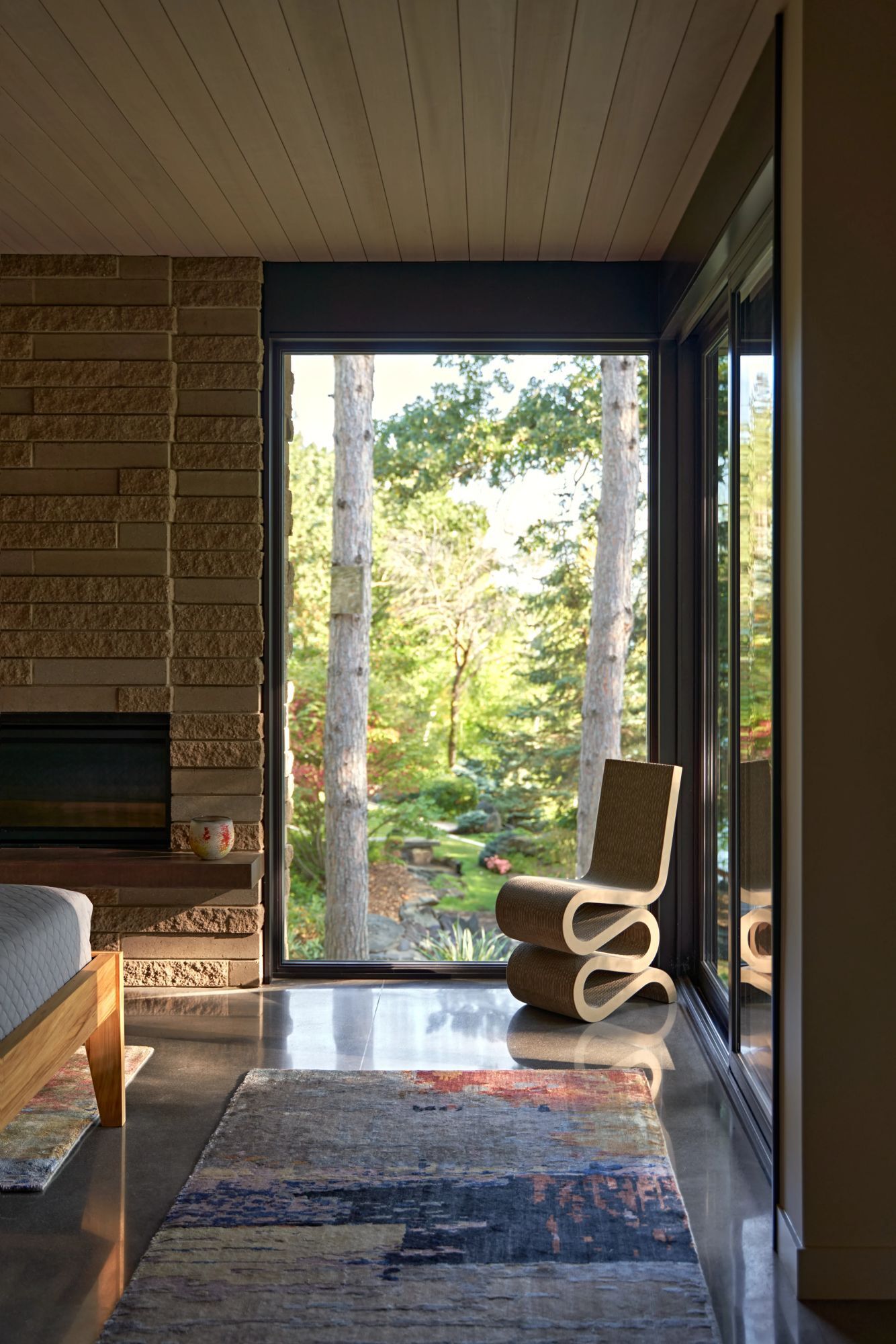 A corner of a modern bedroom featuring a sculptural S-shaped chair beside two full-height windows. The polished concrete floor, patterned rug, wood ceiling, and garden views create a connection between the bedroom and the outdoors.