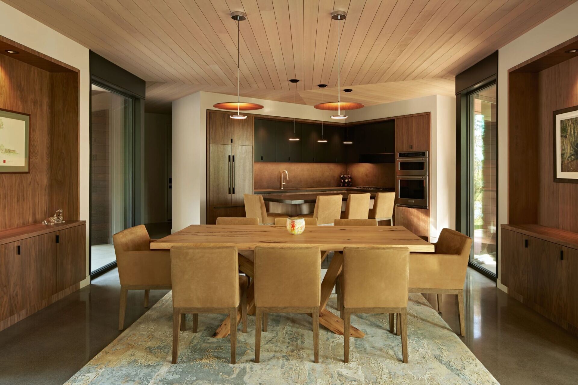 A modern open-plan dining room and kitchen with a large rectangular wood dining table, upholstered tan chairs, dark kitchen cabinets, and a central island. A pale wood ceiling and polished concrete floor extend through the space.