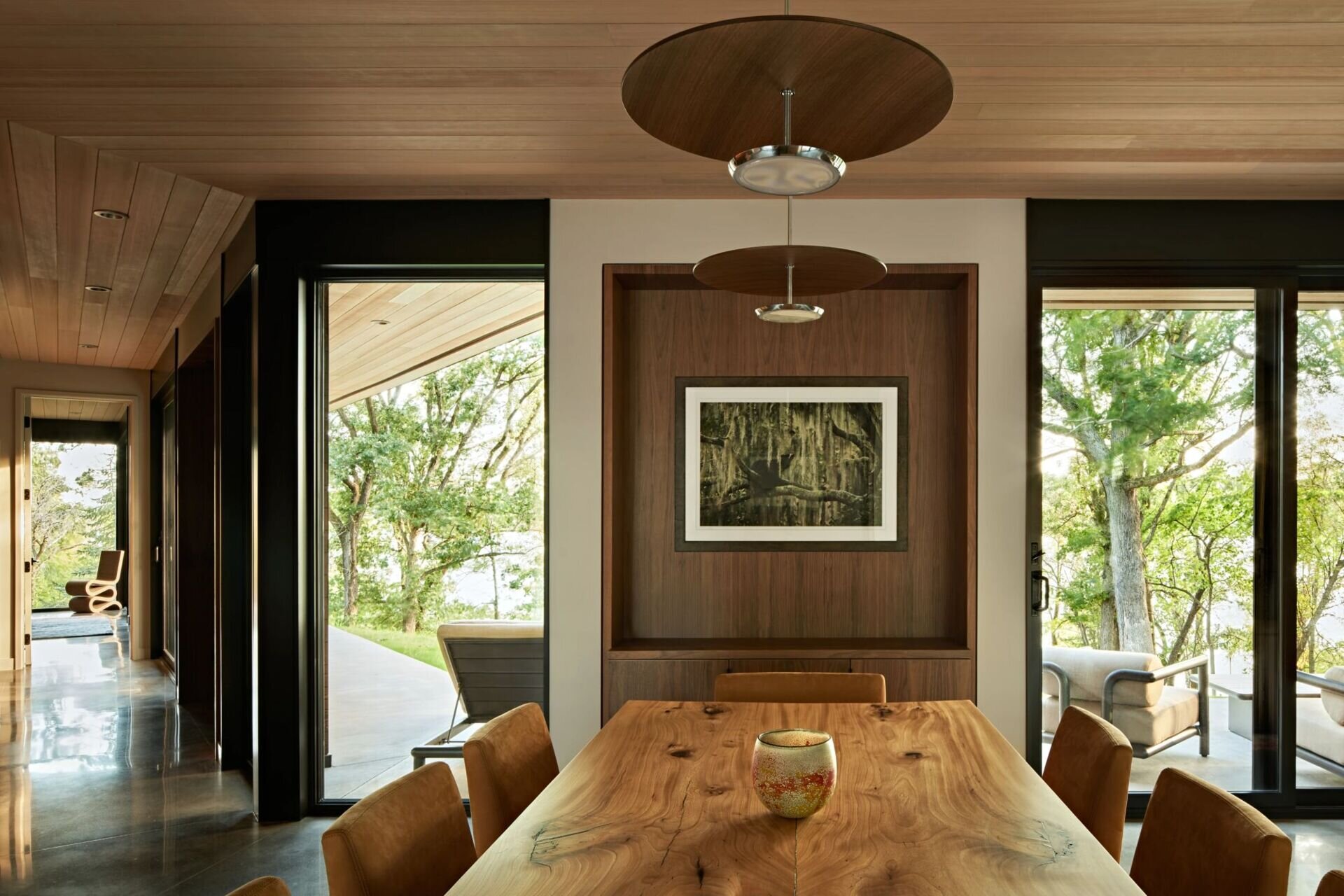 A contemporary dining room featuring a large natural wood table, brown upholstered chairs, and round wood pendant lights. Black-framed glass doors on either side open toward outdoor seating areas and wooded views.