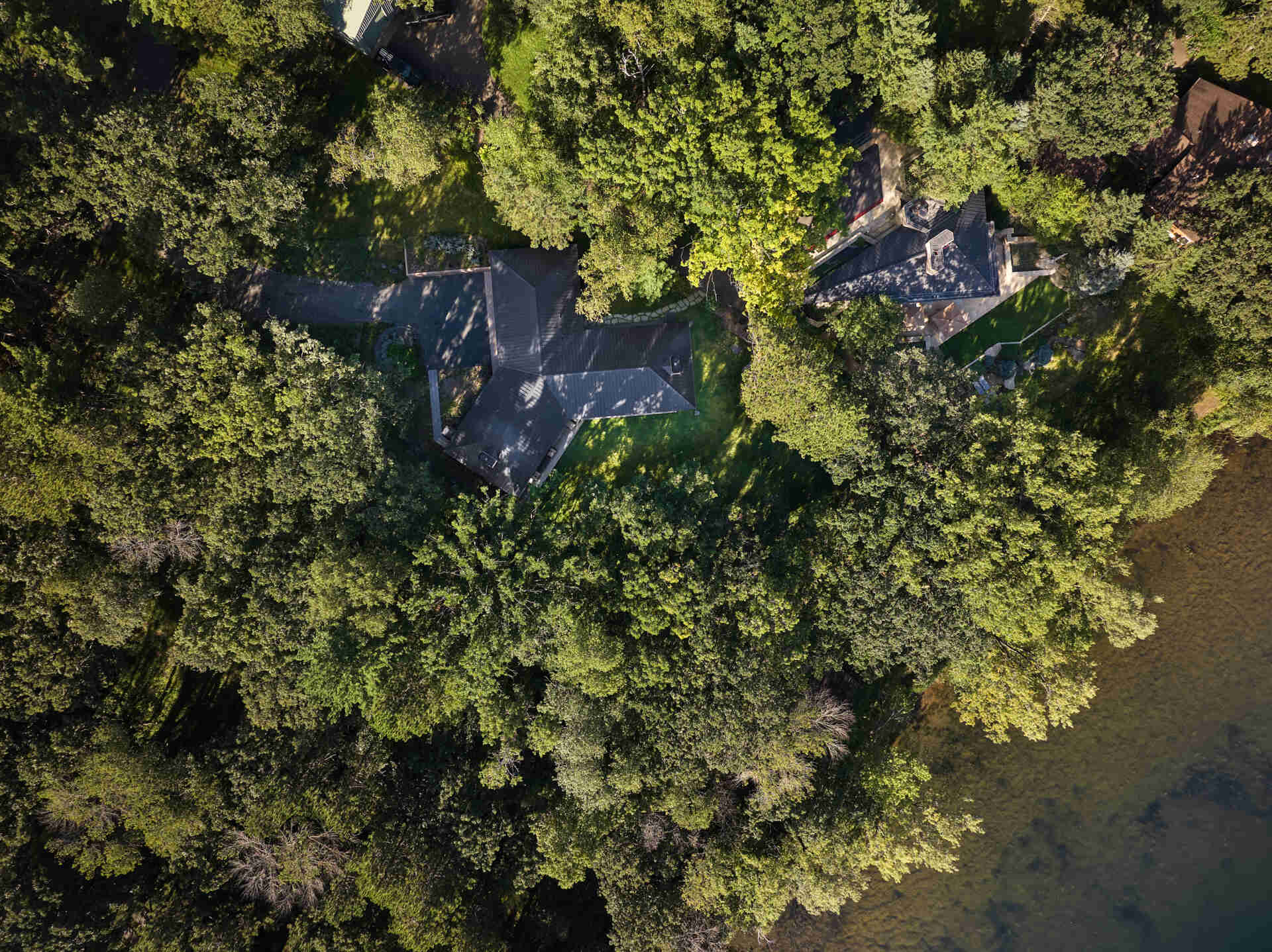 An overhead view of two neighboring lakeside homes in Minnesota, including a Y-shaped modern house and a Frank Lloyd Wright design built from his 1948 plans. Dense trees surround the buildings beside Square Lake.