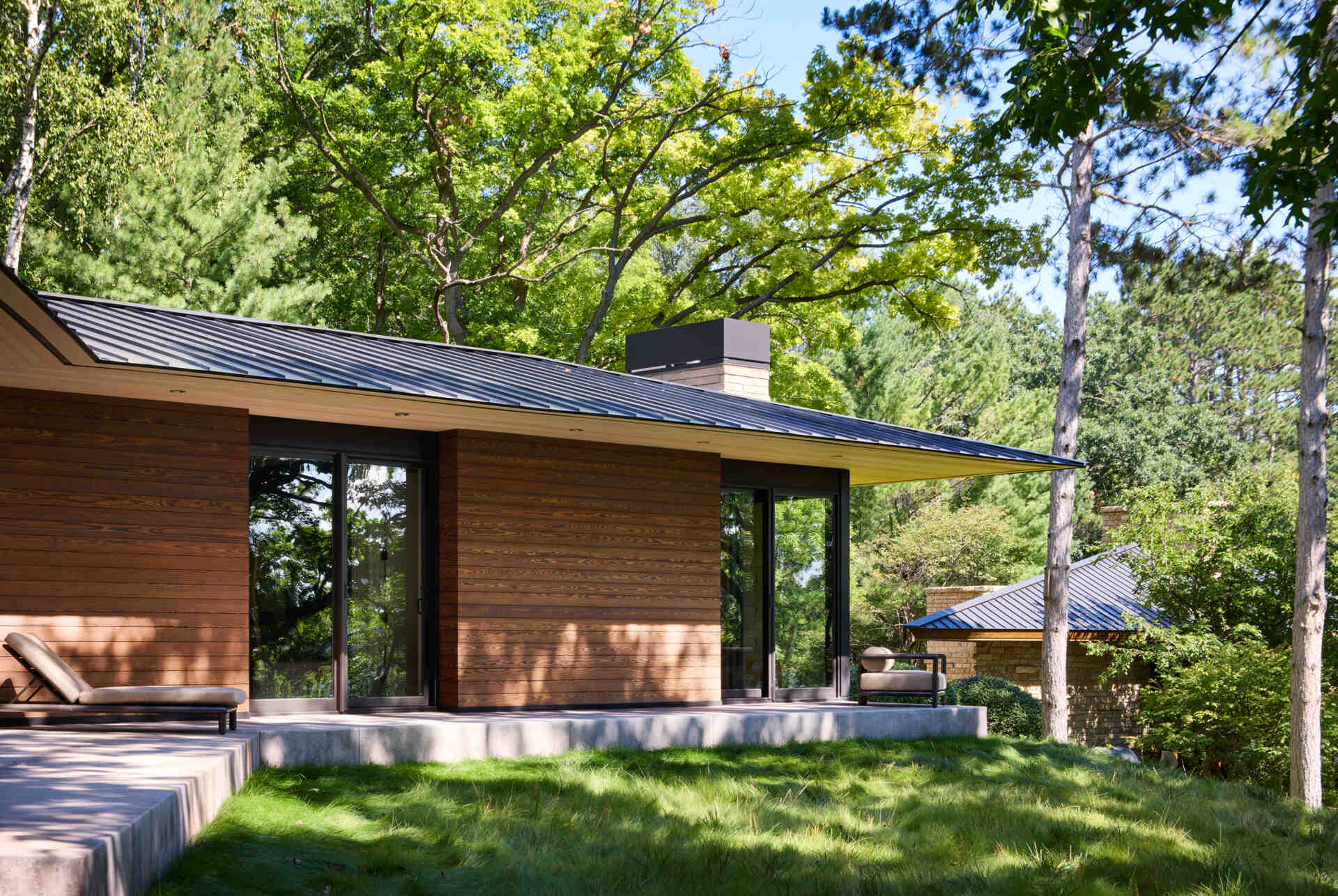 A modern house exterior with horizontal wood siding, black-framed glass doors, and a long metal roof extending over the concrete terrace. Mature trees surround the lawn, with part of the neighboring stone house visible in the background.