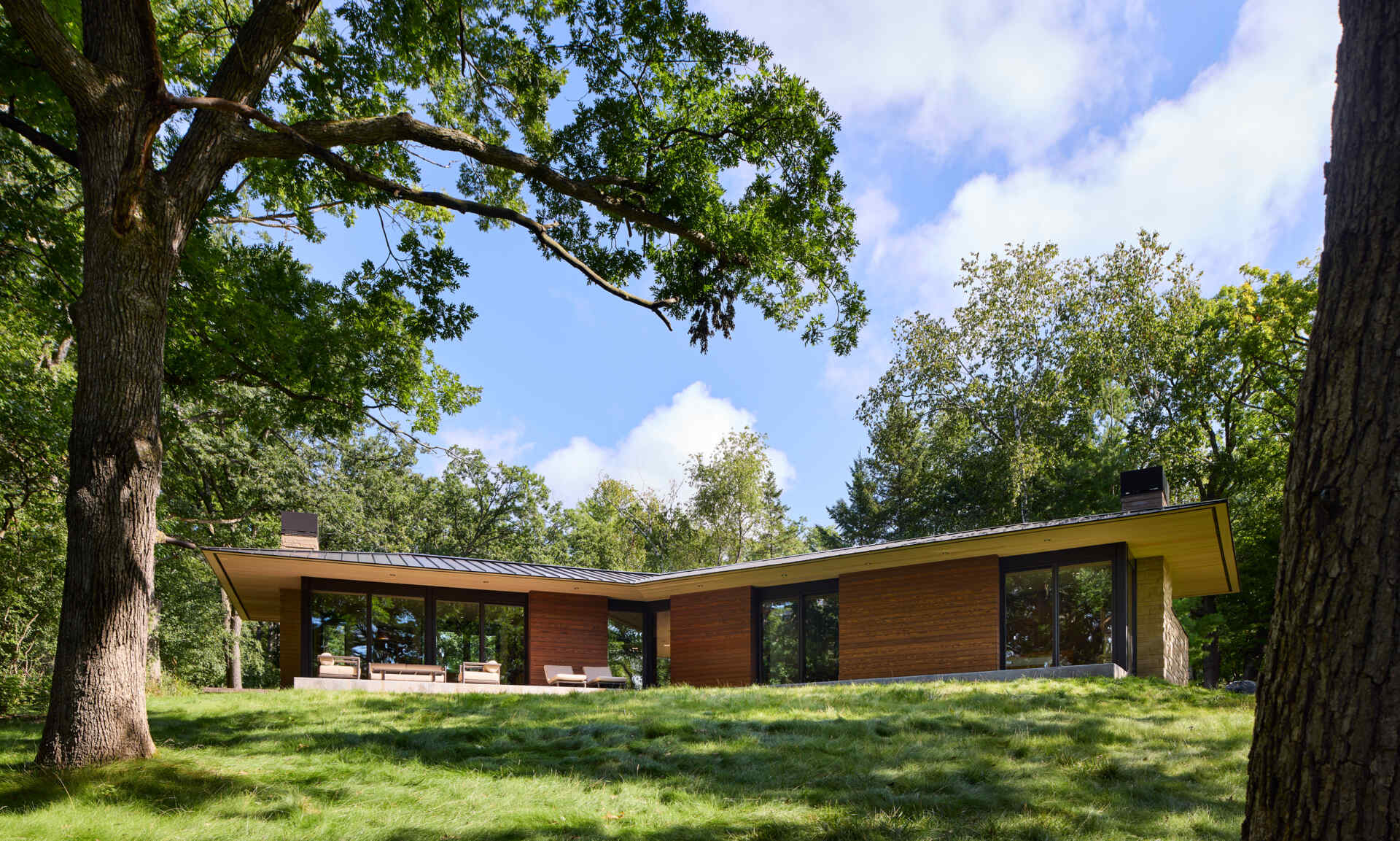 A modern lakeside home with two low roof sections extending in different directions, creating a distinctive angular exterior. Large windows, wood siding, and deep roof overhangs are framed by trees and a grassy lawn.