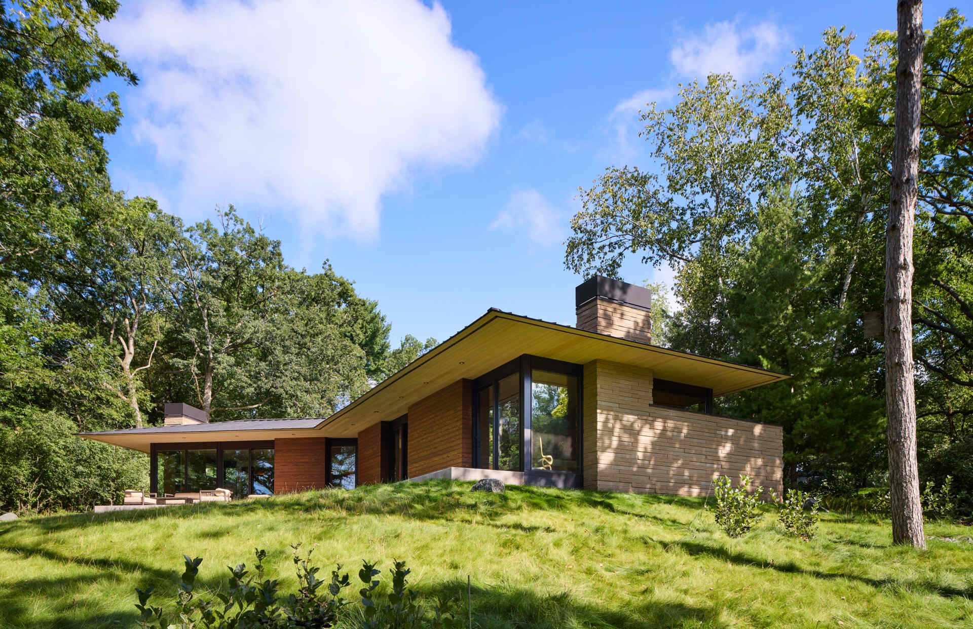 A modern single-story house with a sharply angled roof, horizontal wood siding, pale masonry walls, and large black-framed windows. The home sits on a grassy slope surrounded by mature trees in Minnesota.