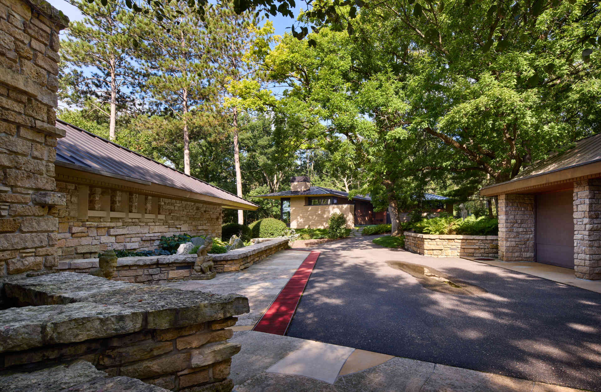 A stone building designed by Frank Lloyd Wright frames the approach toward a neighboring modern home in Minnesota. Limestone walls, sloping roofs, mature trees, and landscaped gardens establish the architectural setting.