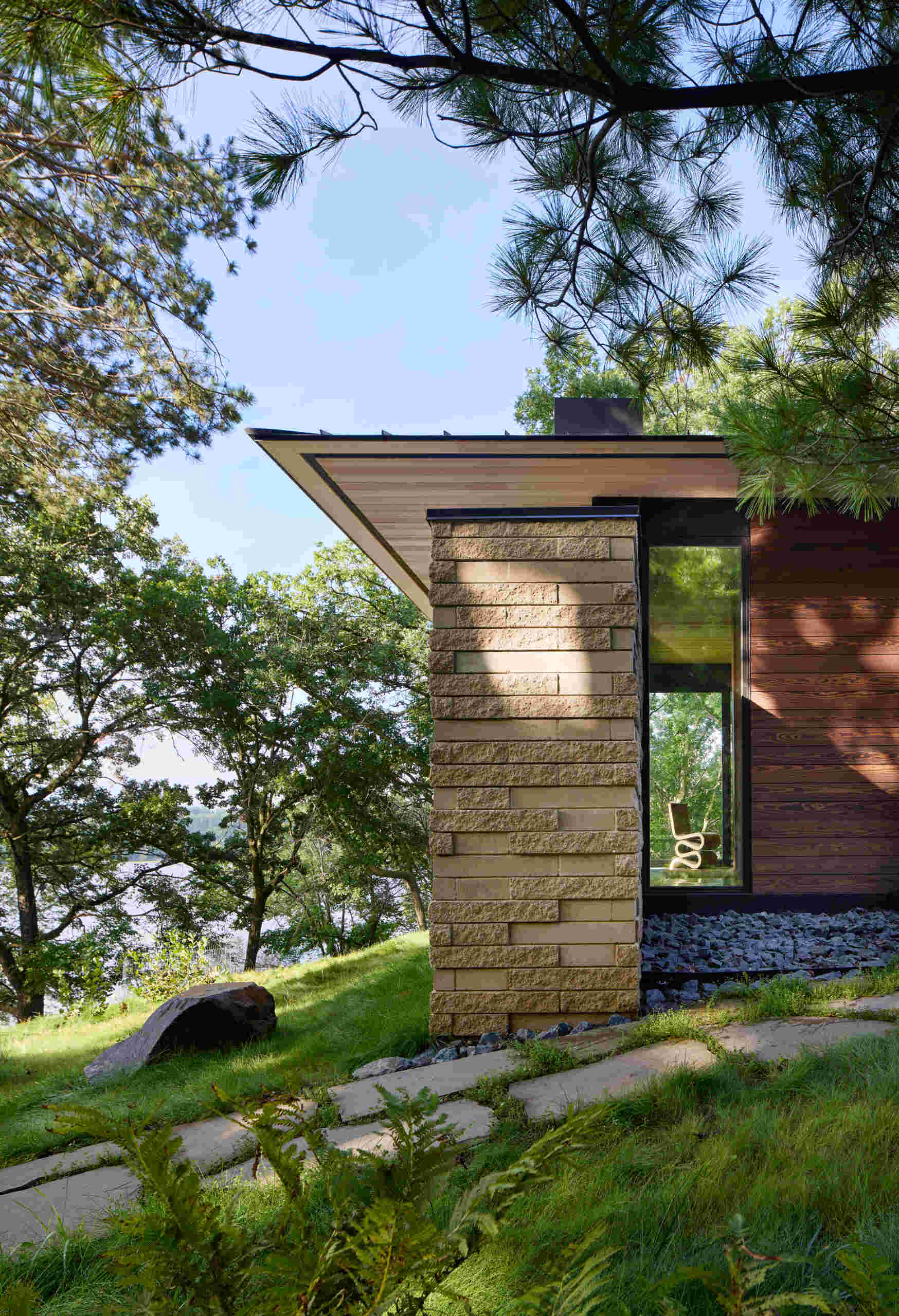 An architectural detail of a modern lakeside house showing a sharply extended roof above a textured stone wall and tall black-framed window. Horizontal wood siding, a wooded hillside, and glimpses of the lake appear around the building.