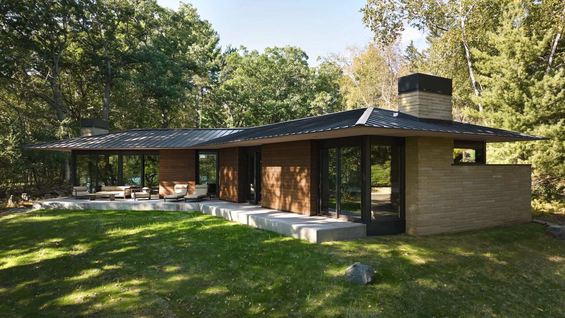 A contemporary single-story house with a dark metal roof, wood-clad walls, floor-to-ceiling glass doors, and a concrete terrace with outdoor seating. The low roof extends beyond the walls to provide shade and shelter.