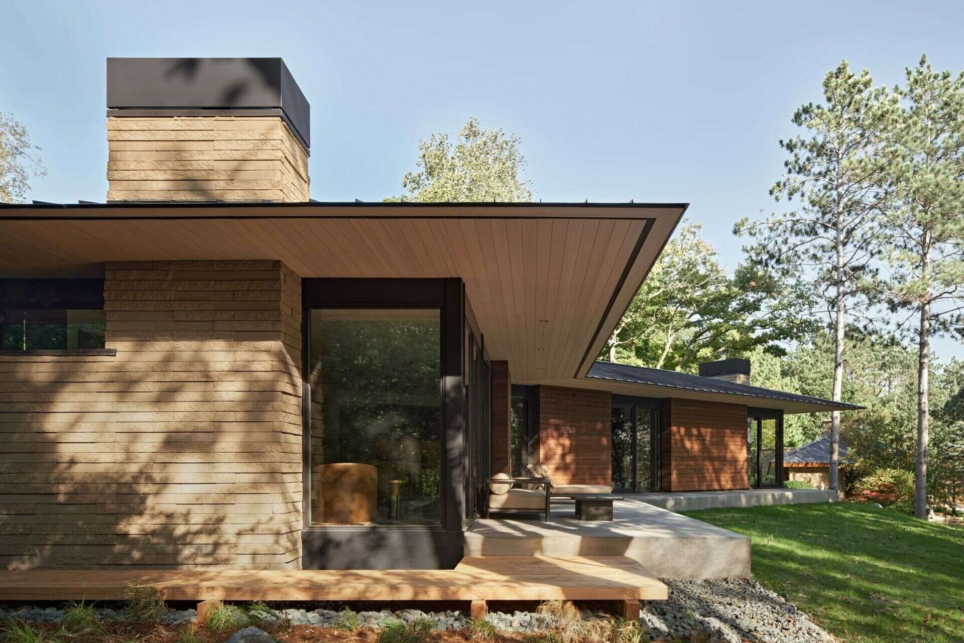 A close exterior view of a modern house featuring a wide cantilevered roof, light-colored masonry, black-framed corner windows, and a wood-lined ceiling beneath the overhang. A concrete patio and wooden walkway extend alongside the building.