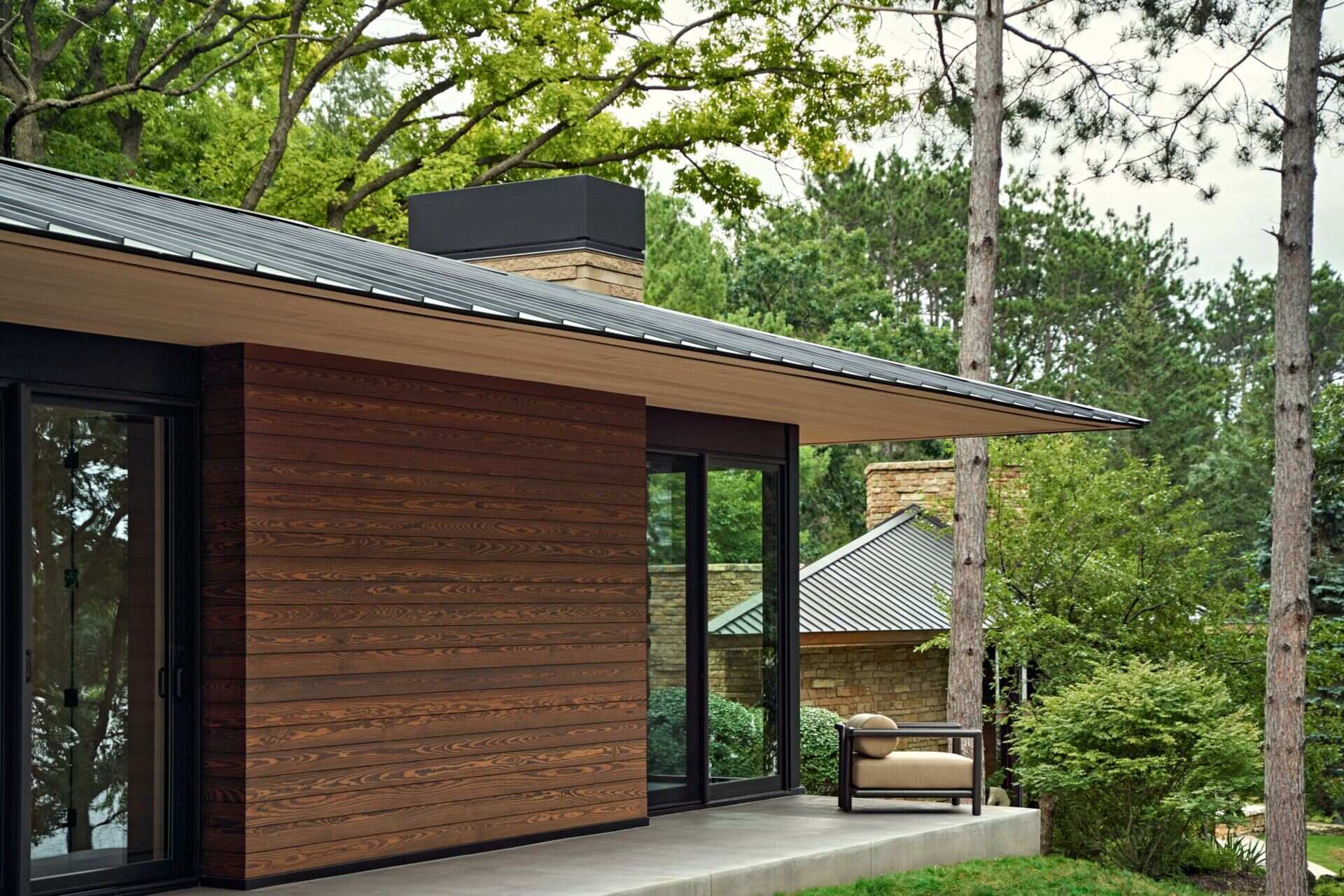 A sheltered concrete terrace along a modern house with horizontal wood siding, large glass doors, and a long metal roof overhang. An outdoor lounge chair sits beside the windows, with a neighboring stone building visible through the trees.