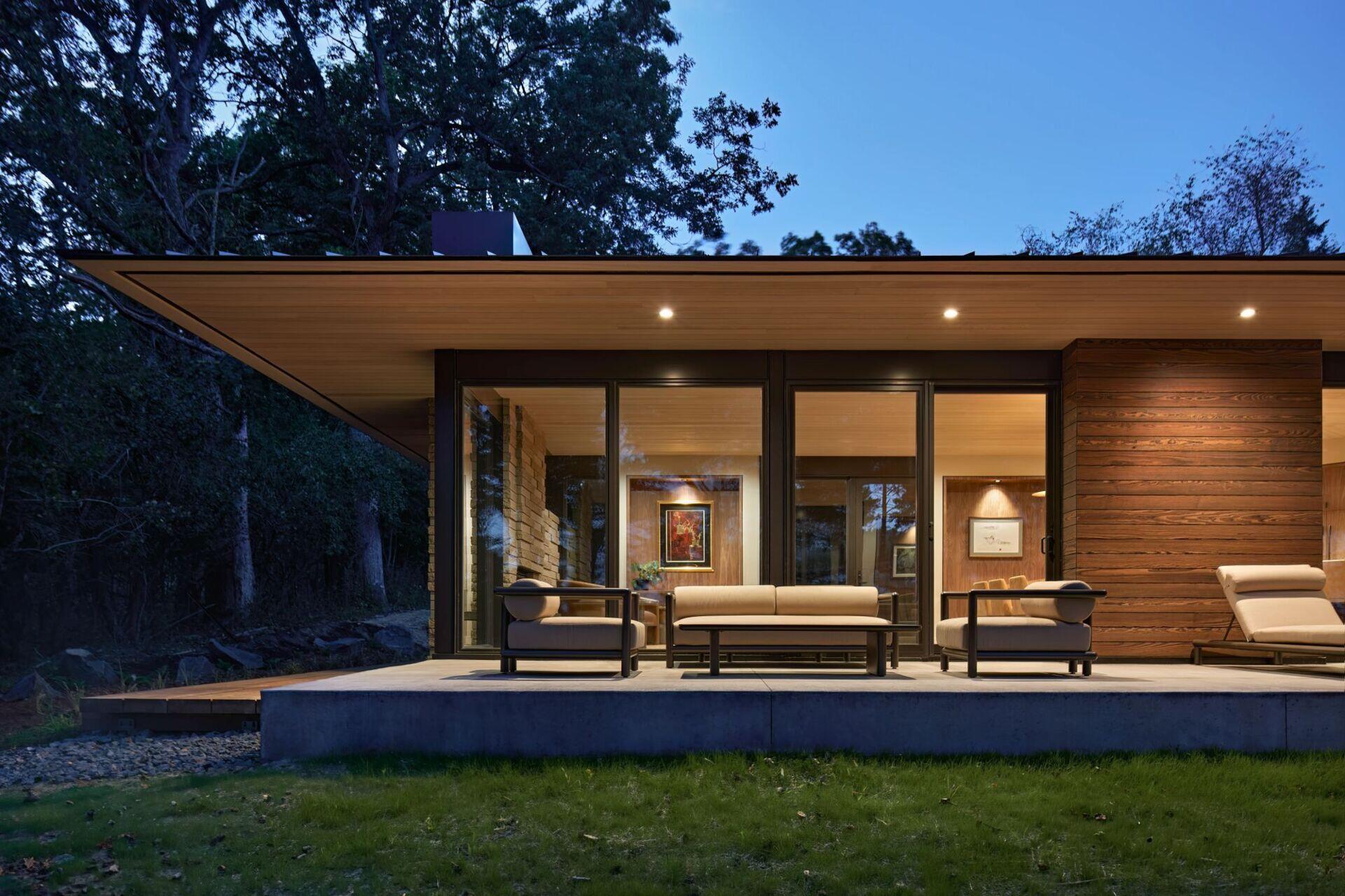 A modern house exterior photographed at dusk, with warm interior lighting visible through large glass doors. Outdoor lounge furniture sits on a concrete terrace beneath a deep wood-lined roof overhang, surrounded by dark woodland.