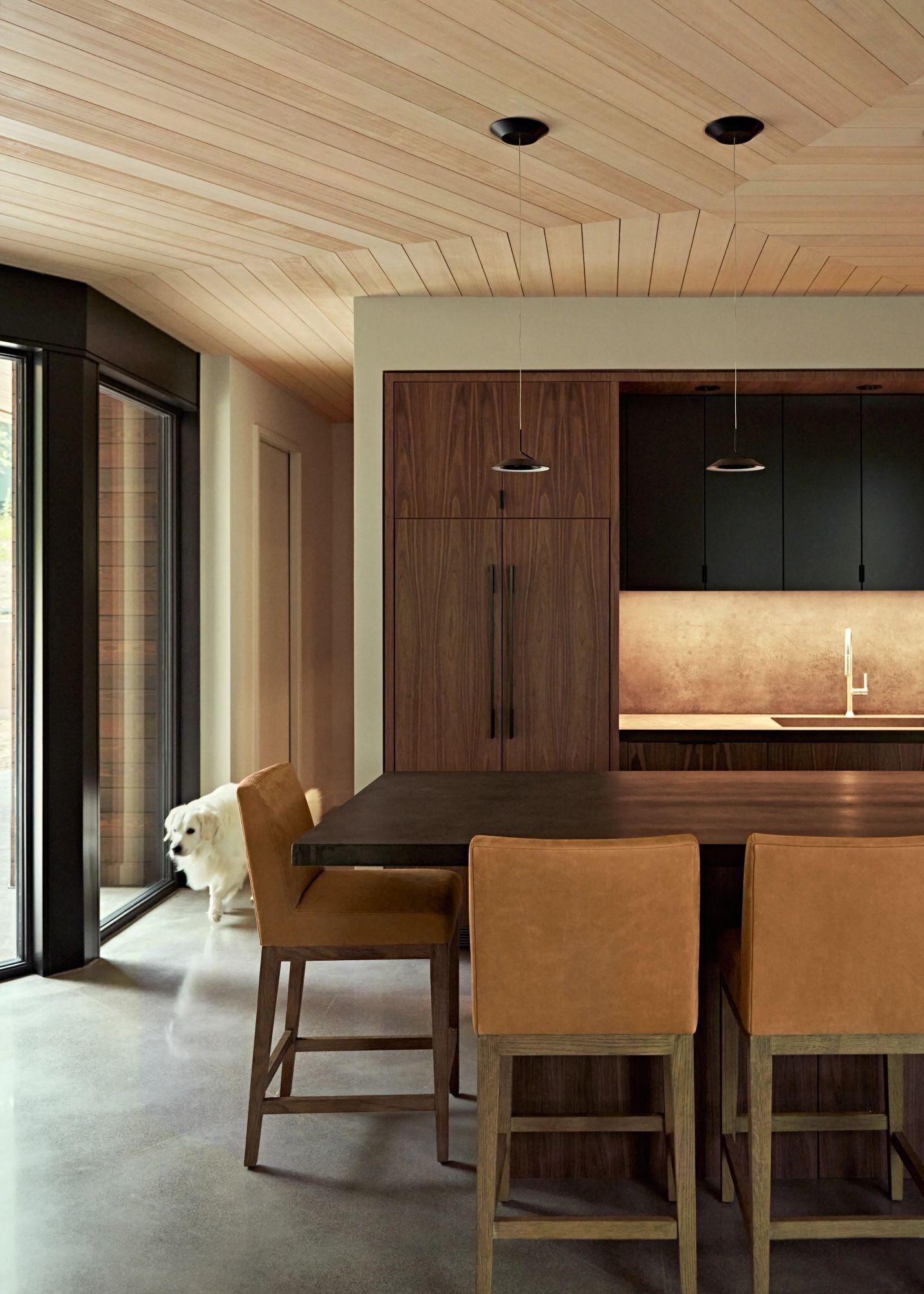 A modern kitchen with dark cabinets, wood-fronted storage, a large island with brown upholstered stools, and a pale wood ceiling that changes direction above the space. Tall black-framed glass doors bring natural light into the room.