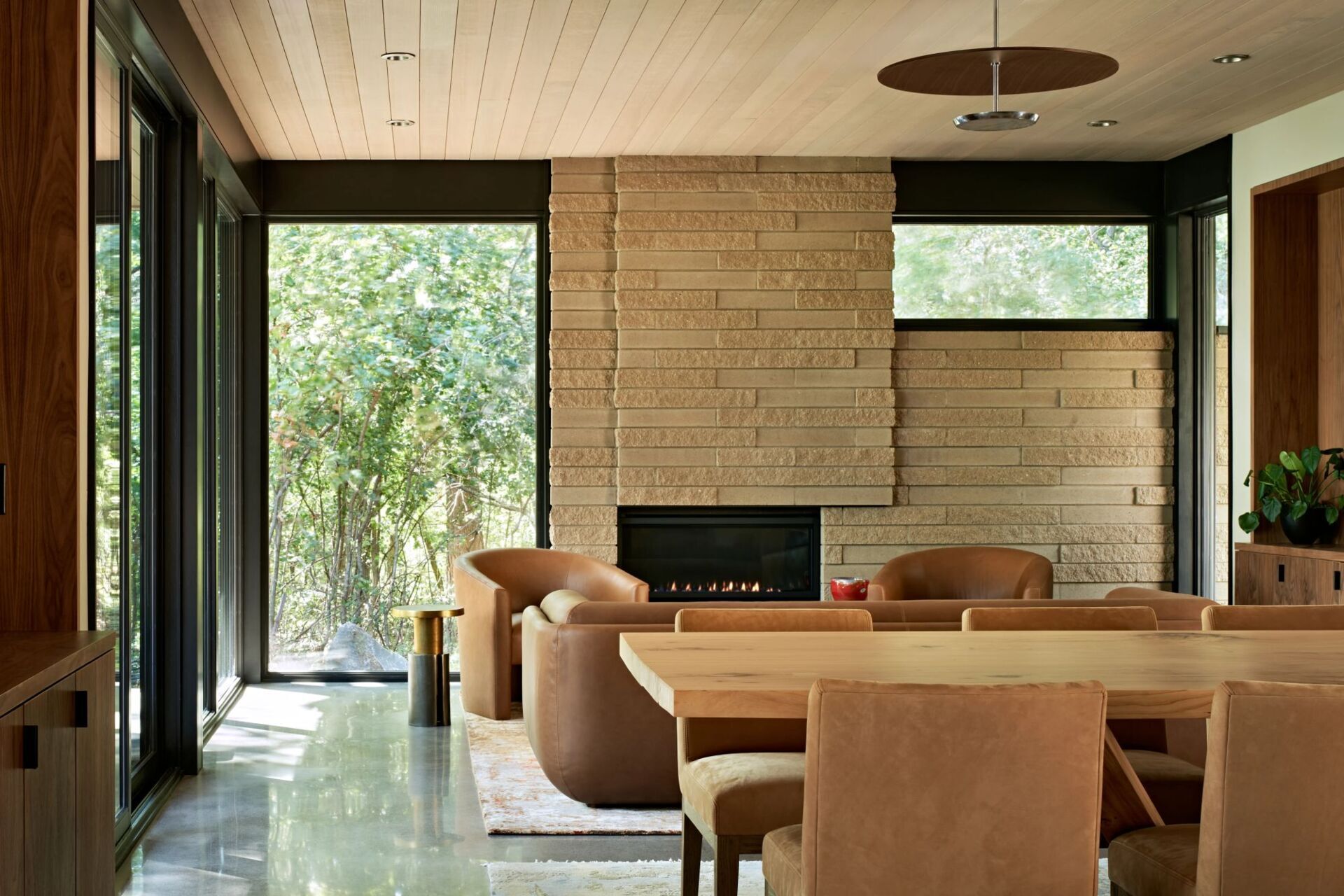 A modern living and dining space with a textured masonry fireplace, curved brown leather seating, floor-to-ceiling windows, and a pale wood ceiling. Polished concrete floors and a large wooden dining table connect the two areas.