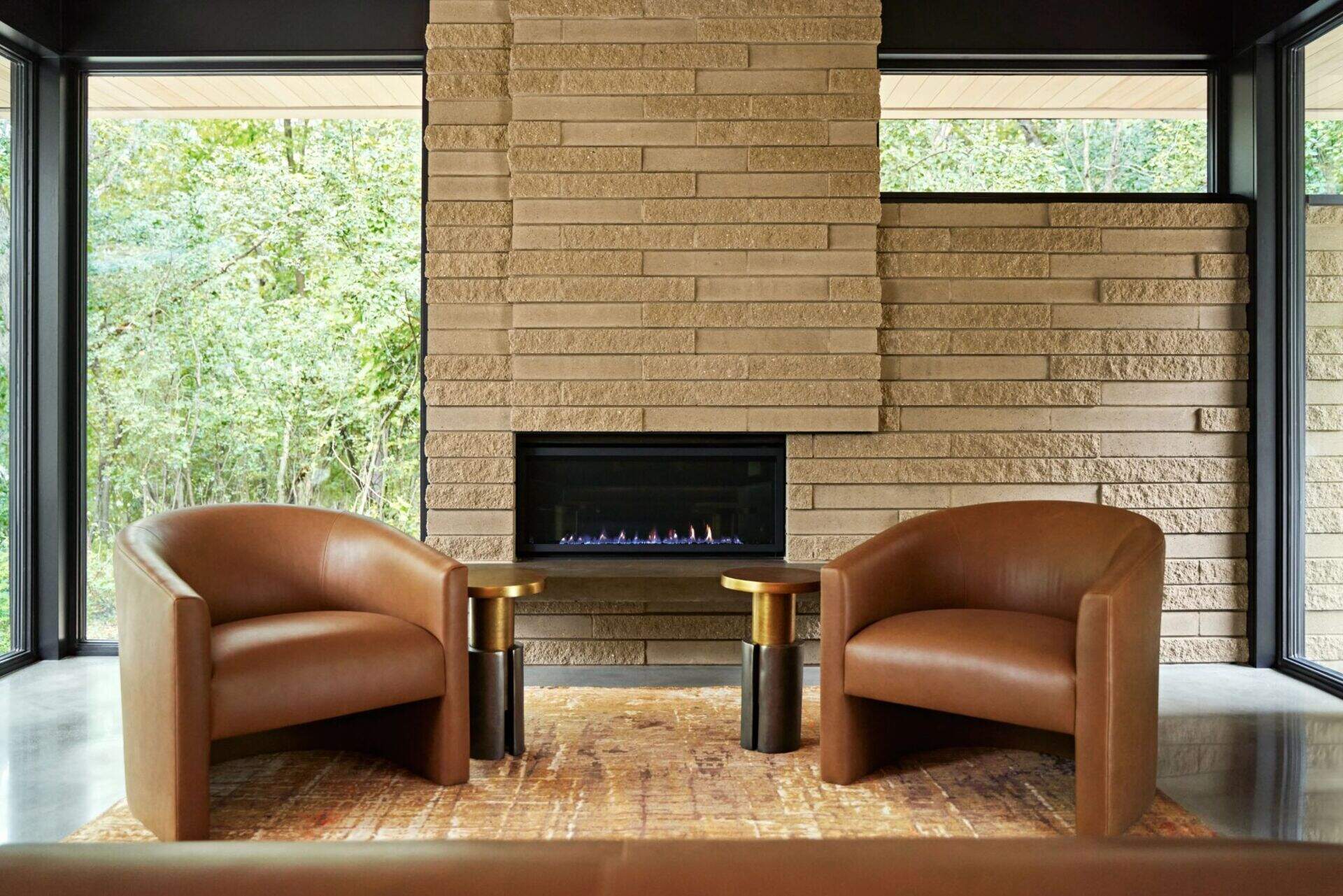 A modern living room fireplace framed by tall windows overlooking the trees. Two curved brown leather armchairs, small brass side tables, and a patterned rug sit in front of the textured horizontal masonry wall.