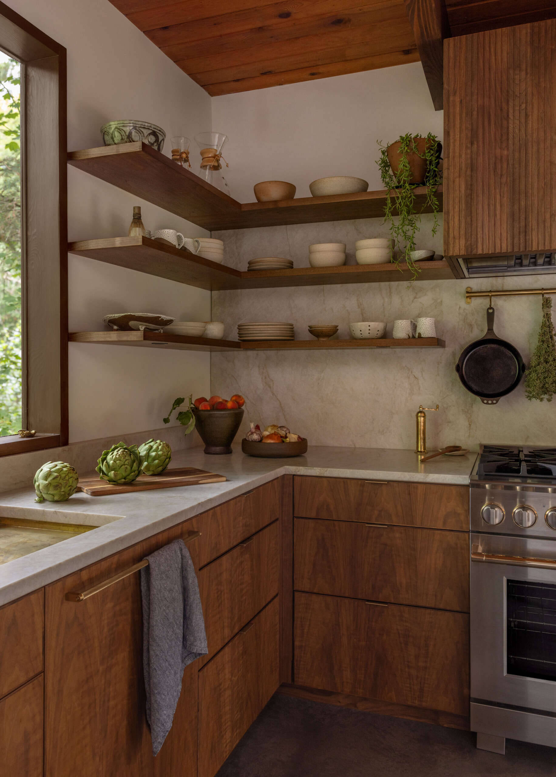 Explore the renovated kitchen of The Burrows, a mid-century modern home in Seattle by chadbourne + doss architects. Custom walnut cabinetry, slatted wood details, brass fixtures, pale stone countertops, a brass chef's sink, and a sculptural kitchen island bring warmth and functionality to the space.