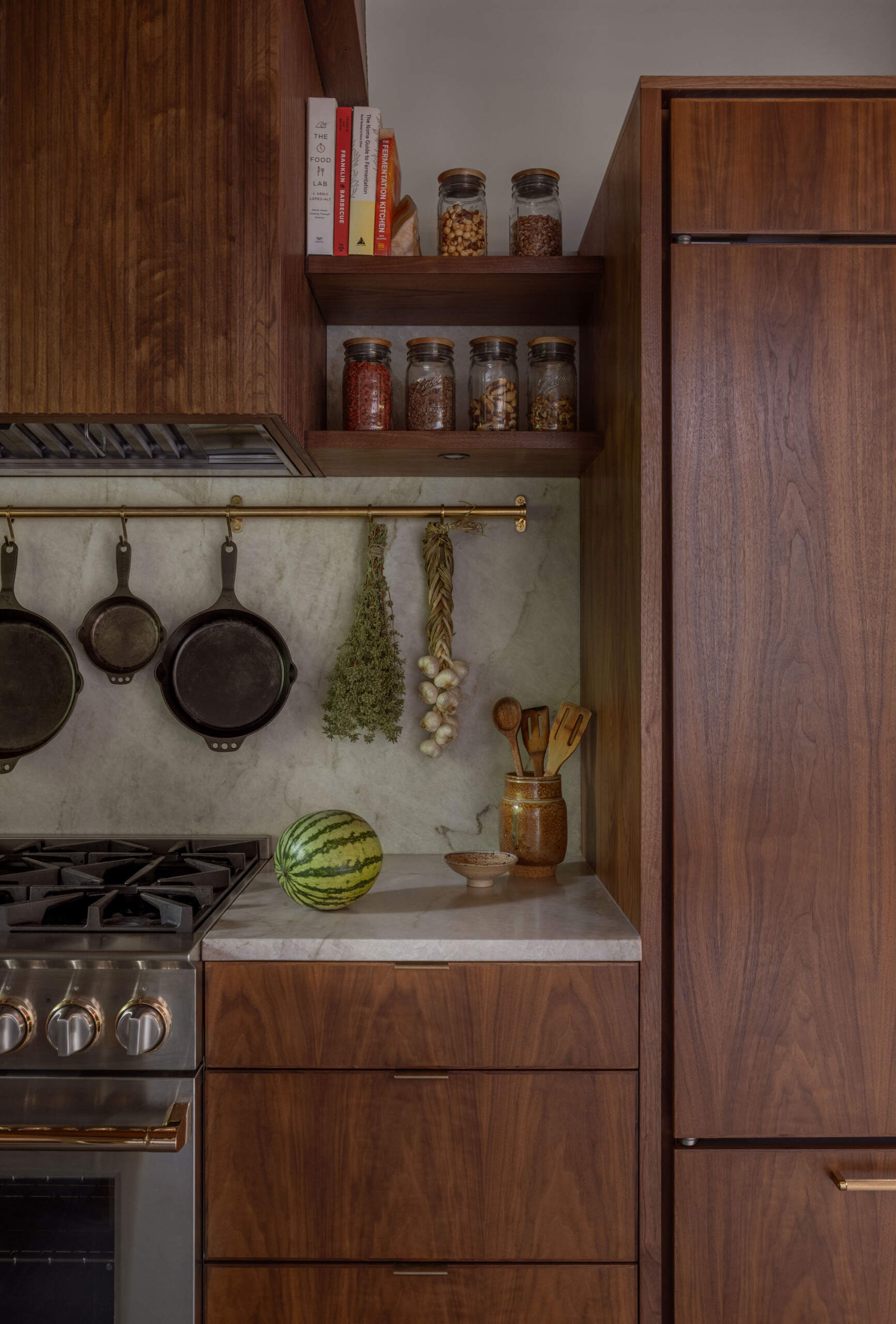 Explore the renovated kitchen of The Burrows, a mid-century modern home in Seattle by chadbourne + doss architects. Custom walnut cabinetry, slatted wood details, brass fixtures, pale stone countertops, a brass chef's sink, and a sculptural kitchen island bring warmth and functionality to the space.