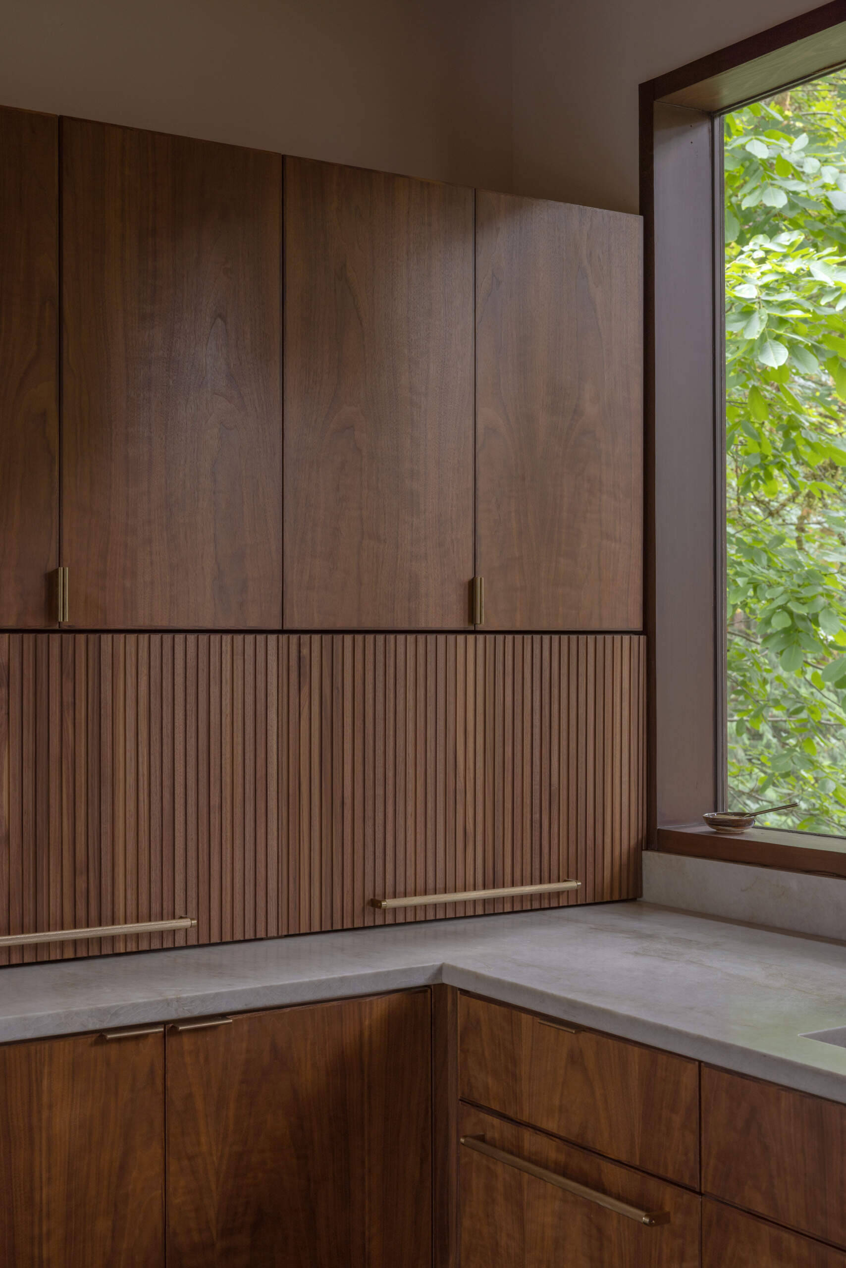 Explore the renovated kitchen of The Burrows, a mid-century modern home in Seattle by chadbourne + doss architects. Custom walnut cabinetry, slatted wood details, brass fixtures, pale stone countertops, a brass chef's sink, and a sculptural kitchen island bring warmth and functionality to the space.