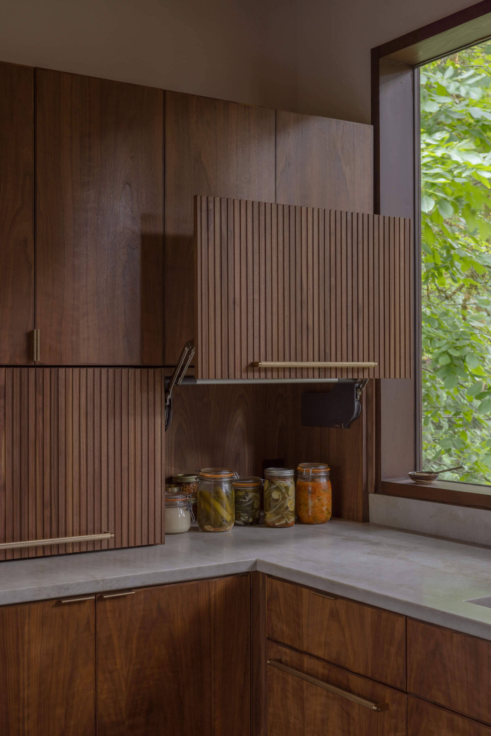 Explore the renovated kitchen of The Burrows, a mid-century modern home in Seattle by chadbourne + doss architects. Custom walnut cabinetry, slatted wood details, brass fixtures, pale stone countertops, a brass chef's sink, and a sculptural kitchen island bring warmth and functionality to the space.