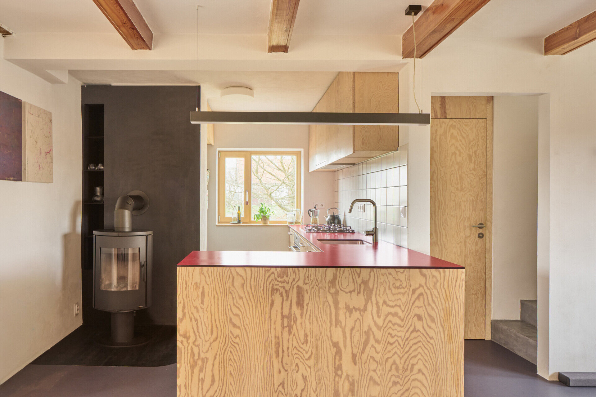 The redesigned kitchen and dining area in this Ne?ín home by Facha architekti combines custom pine plywood cabinetry, natural linoleum flooring and a rotating wood-burning stove within the reworked ground floor.