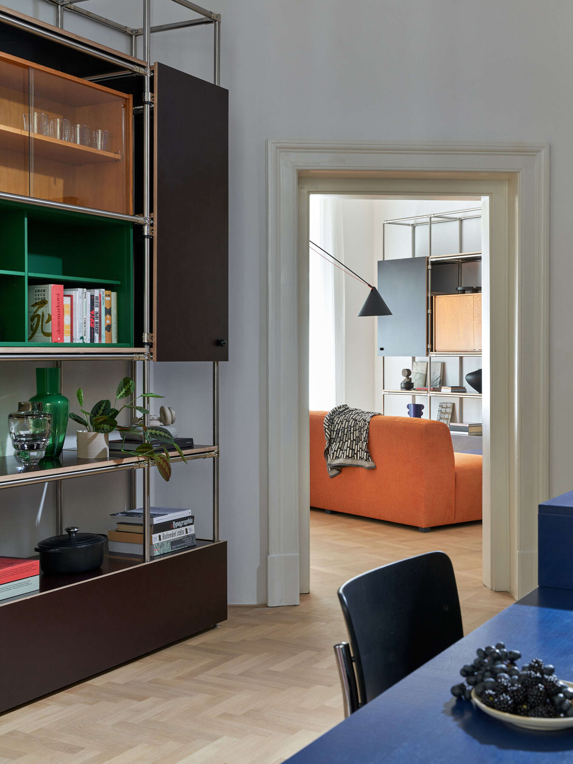 A view through a historic panelled doorway reveals an orange sofa and open shelving in a modern living room. The adjoining space includes a deep blue kitchen island and pale herringbone flooring.
