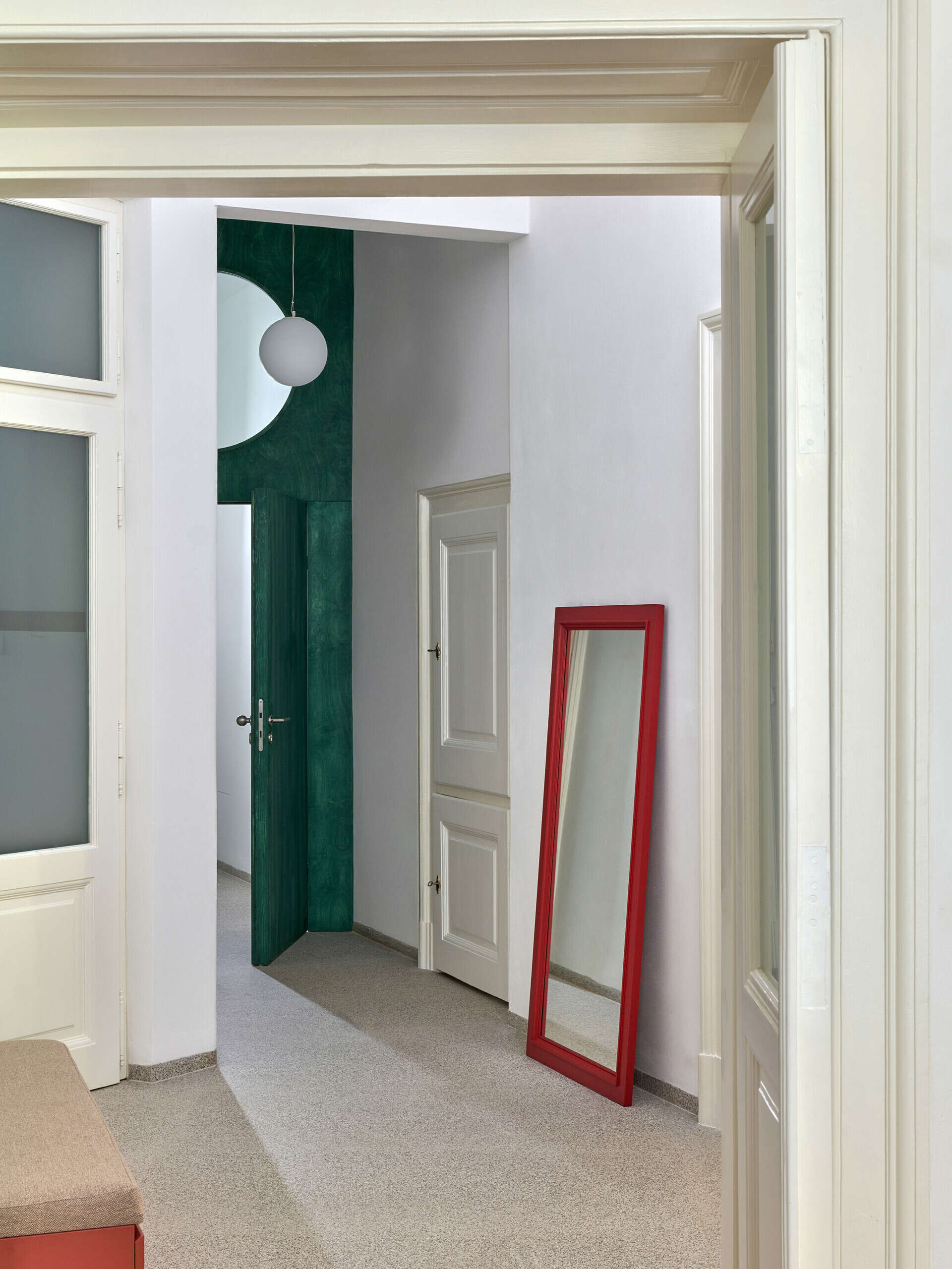 A renovated Prague apartment hallway combines original white panelled door frames with a dark green door, red-framed floor mirror, and pale speckled flooring.