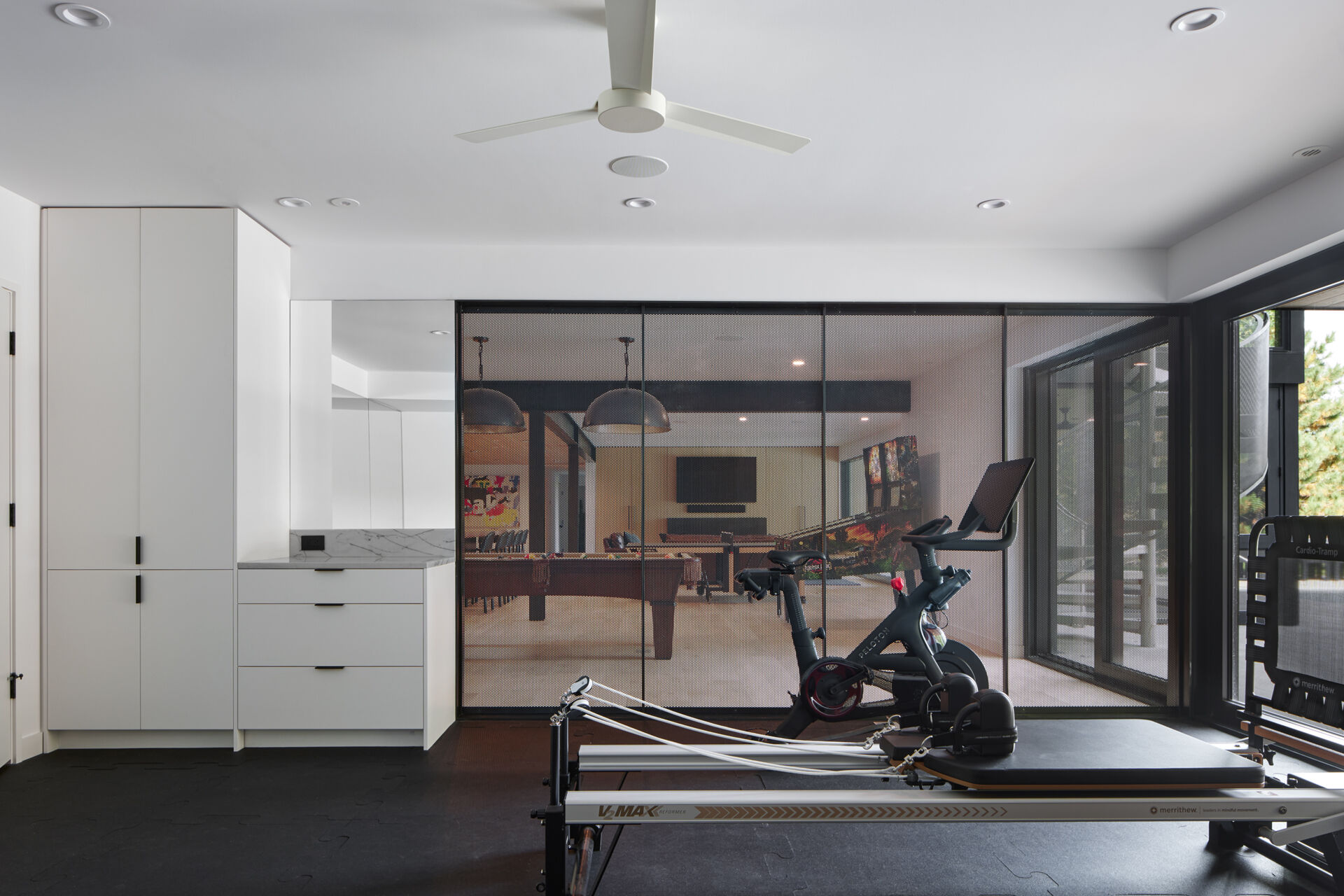 A modern home gym with an exercise bike, Pilates equipment, white storage cabinets, and large windows. A black mesh partition separates the workout area from the adjoining games room.