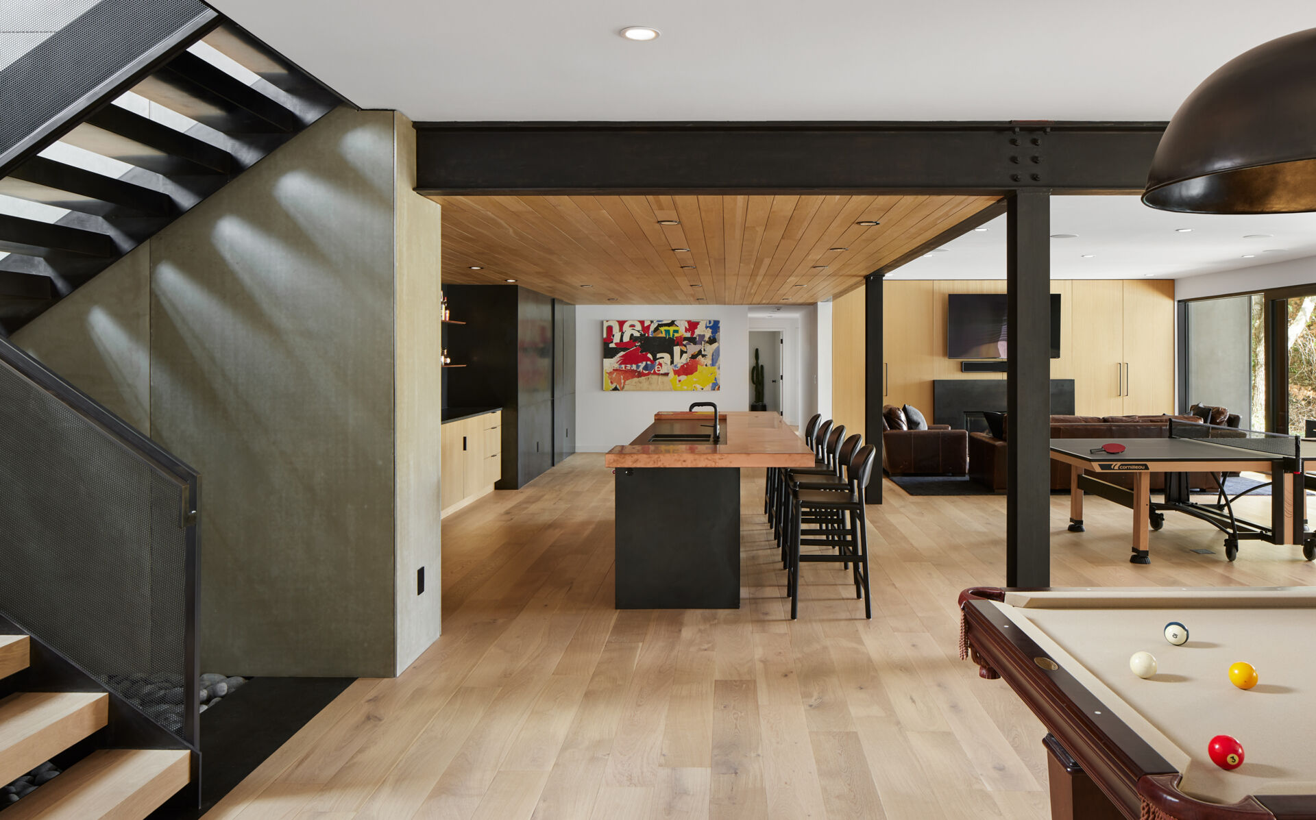 A modern basement entertainment area with a pool table, table tennis table, lounge, and long bar counter. Exposed black steel beams, light wood floors, and a wood ceiling bring the different recreation spaces together.
