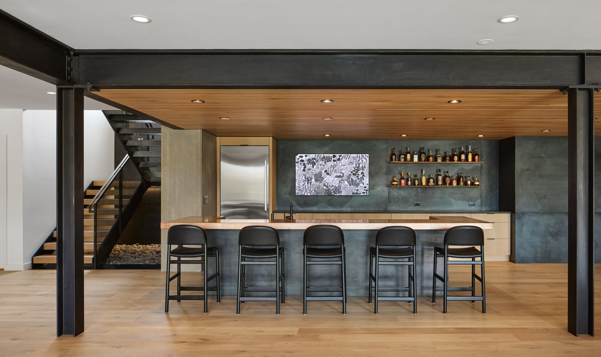 A contemporary basement bar featuring a long counter, five black bar stools, open bottle shelving, and a dark gray wall. Exposed steel beams and a wood ceiling add an industrial touch to the entertainment space.