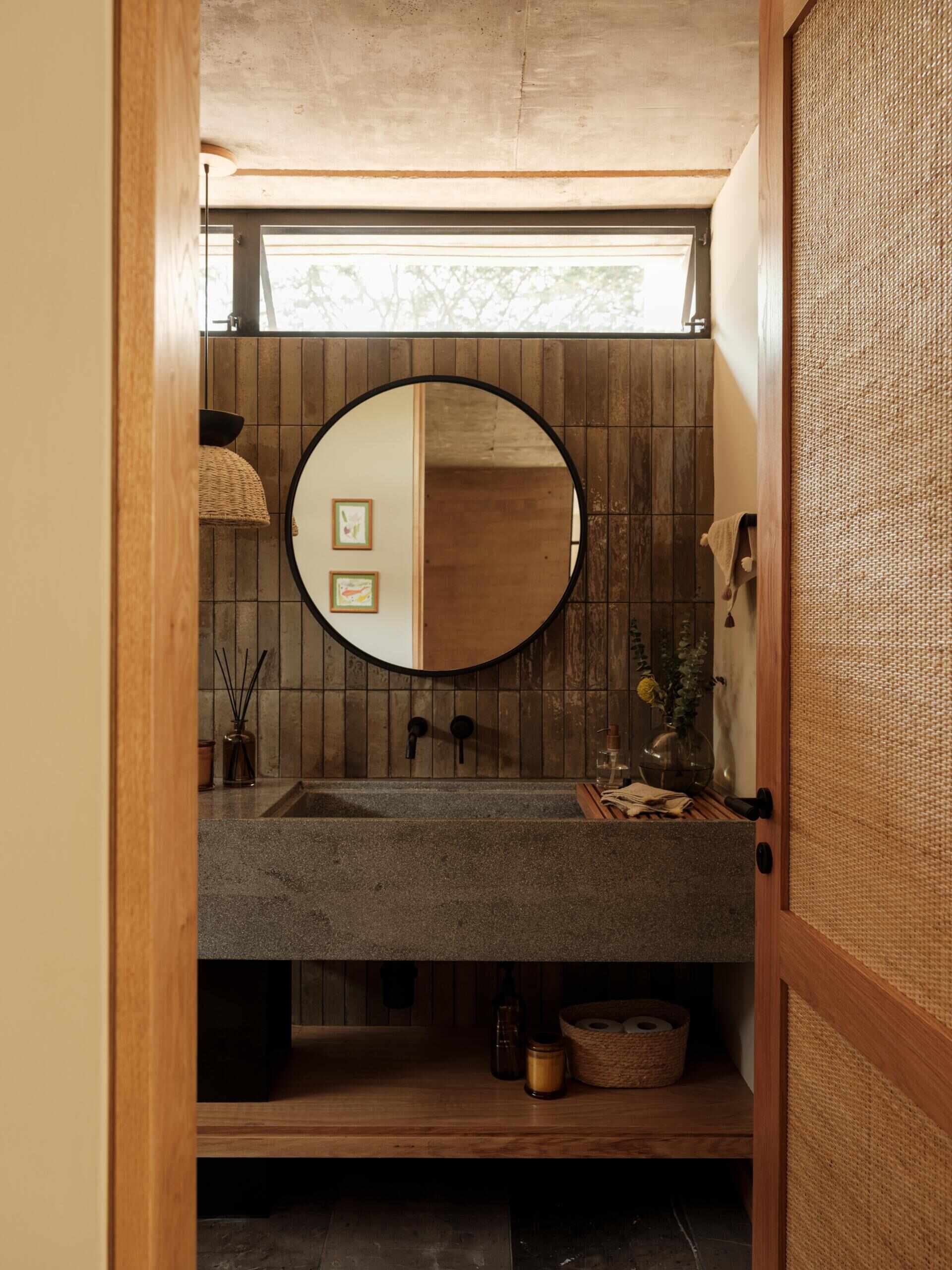 Explore a modern bathroom featuring earthy vertical tiles, a stone vanity with an integrated sink, matte black fixtures, a round mirror, and warm wood details. A beautiful example of natural materials in contemporary bathroom design.