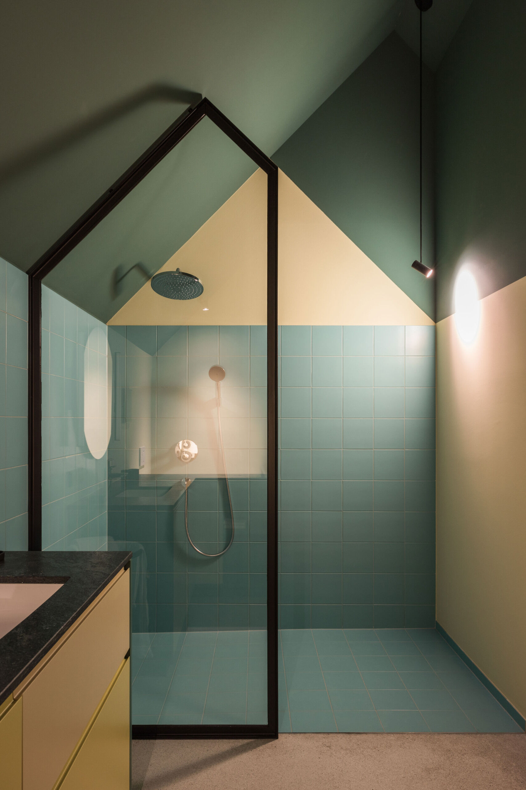 A colourful contemporary bathroom beneath a pitched roof features turquoise shower tiles, an unusual house-shaped glass enclosure, green walls, and geometric architectural details.