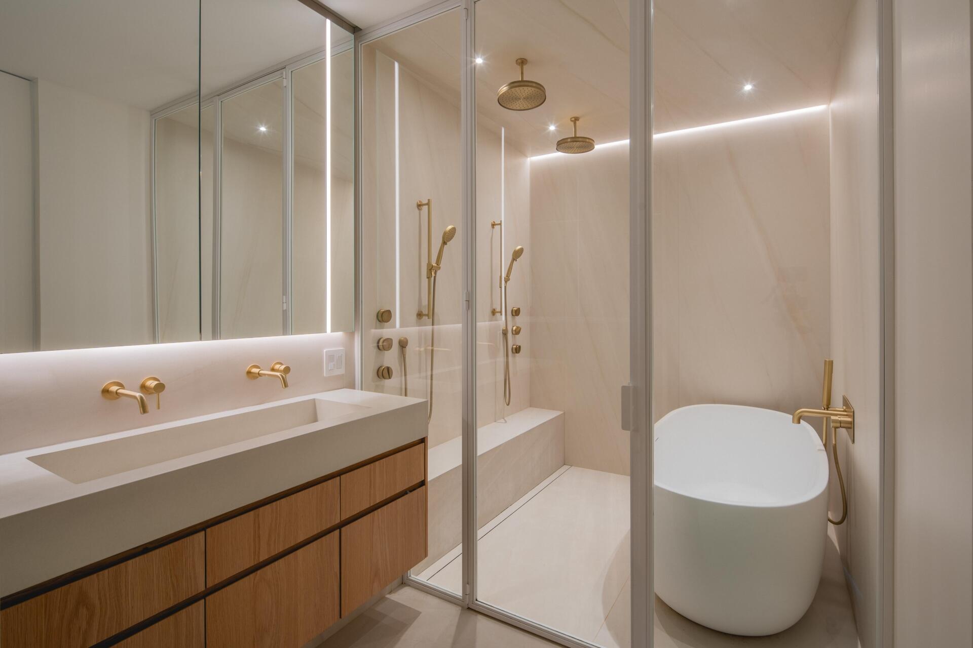 Explore the modern bathroom at The Lens by MU Architecture, featuring beige porcelain, brushed brass fixtures, a floating wood vanity, glass-enclosed shower, freestanding bathtub, and soft integrated lighting.
